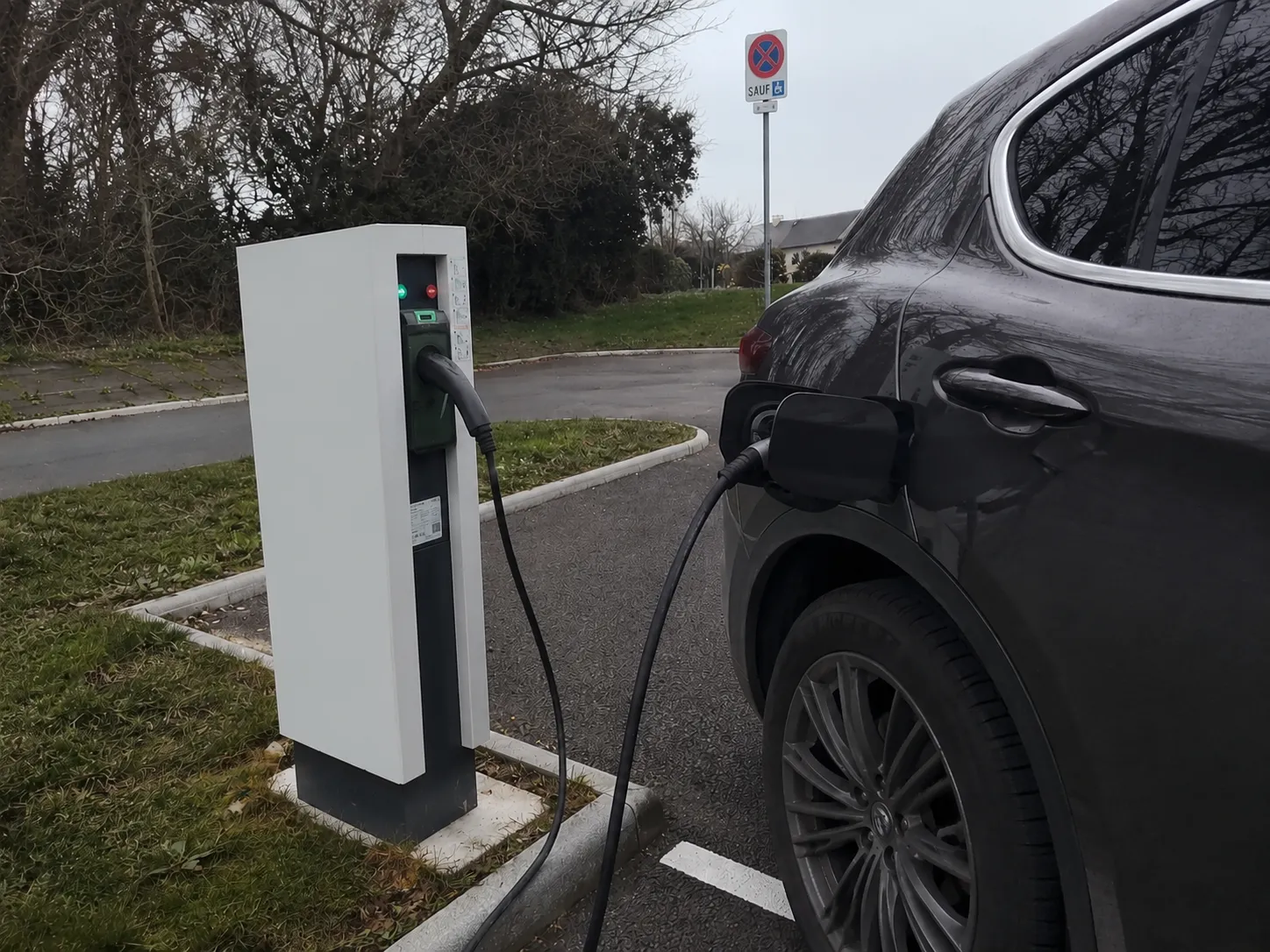 Une voiture électrique noire en train de se charger sur une borne de recharge dans un parking, avec un panneau indiquant une interdiction de stationner sauf pour personnes handicapées en arrière-plan.