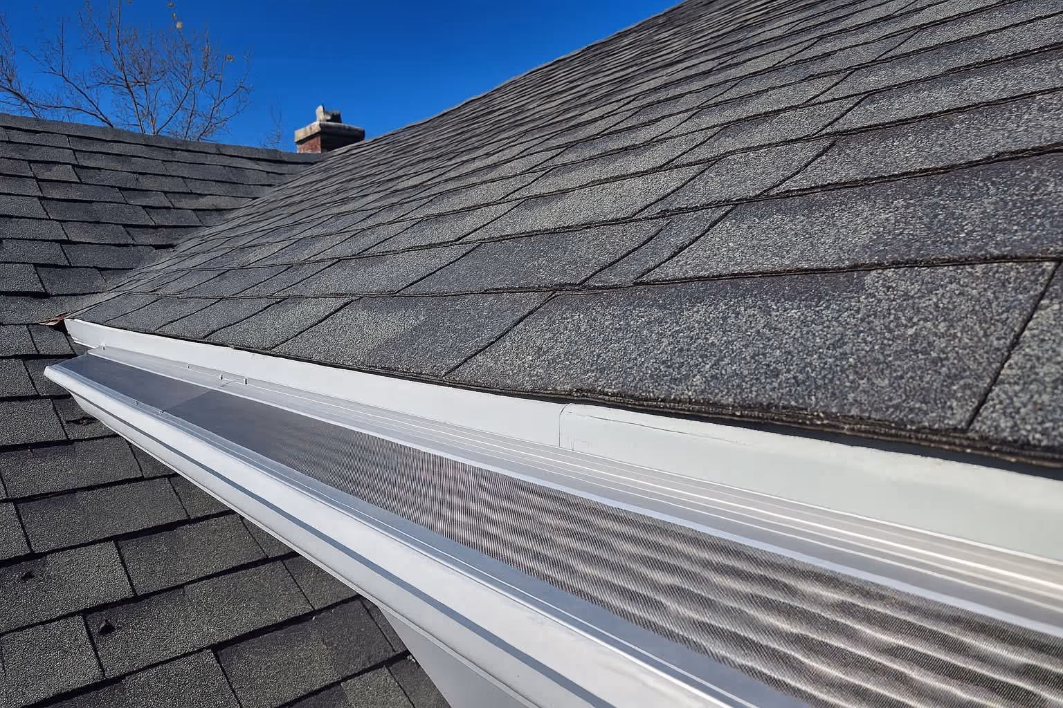 Roof with gray shingles and white gutter protection installed under a clear blue sky.