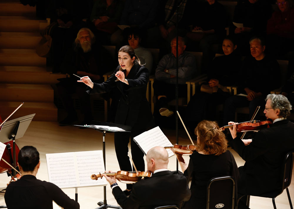 A conductor gestures to a group of string musicians performing on stage. Photo by Winslow Townson.