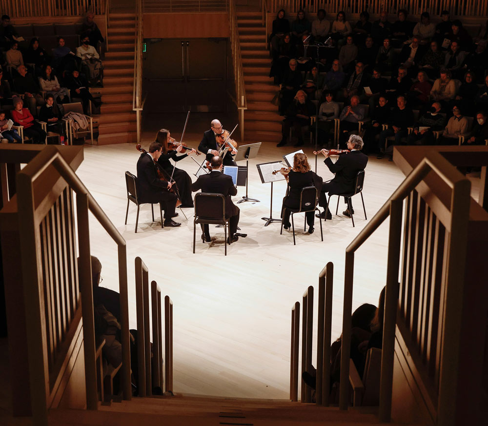 A group of string musicians sit in a circle facing each other on stage.Photo by Winslow Townson.