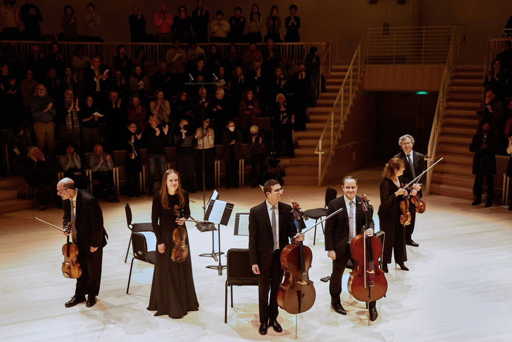 A group of string musicians stand on stage in a circle facing towards the audience.Photo by Winslow Townson.