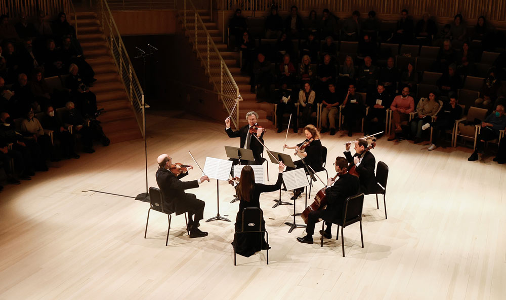 A group of five string musicians sitting and a standing conductor form a circle on stage facing towards each other. Photo by Winslow Townson.