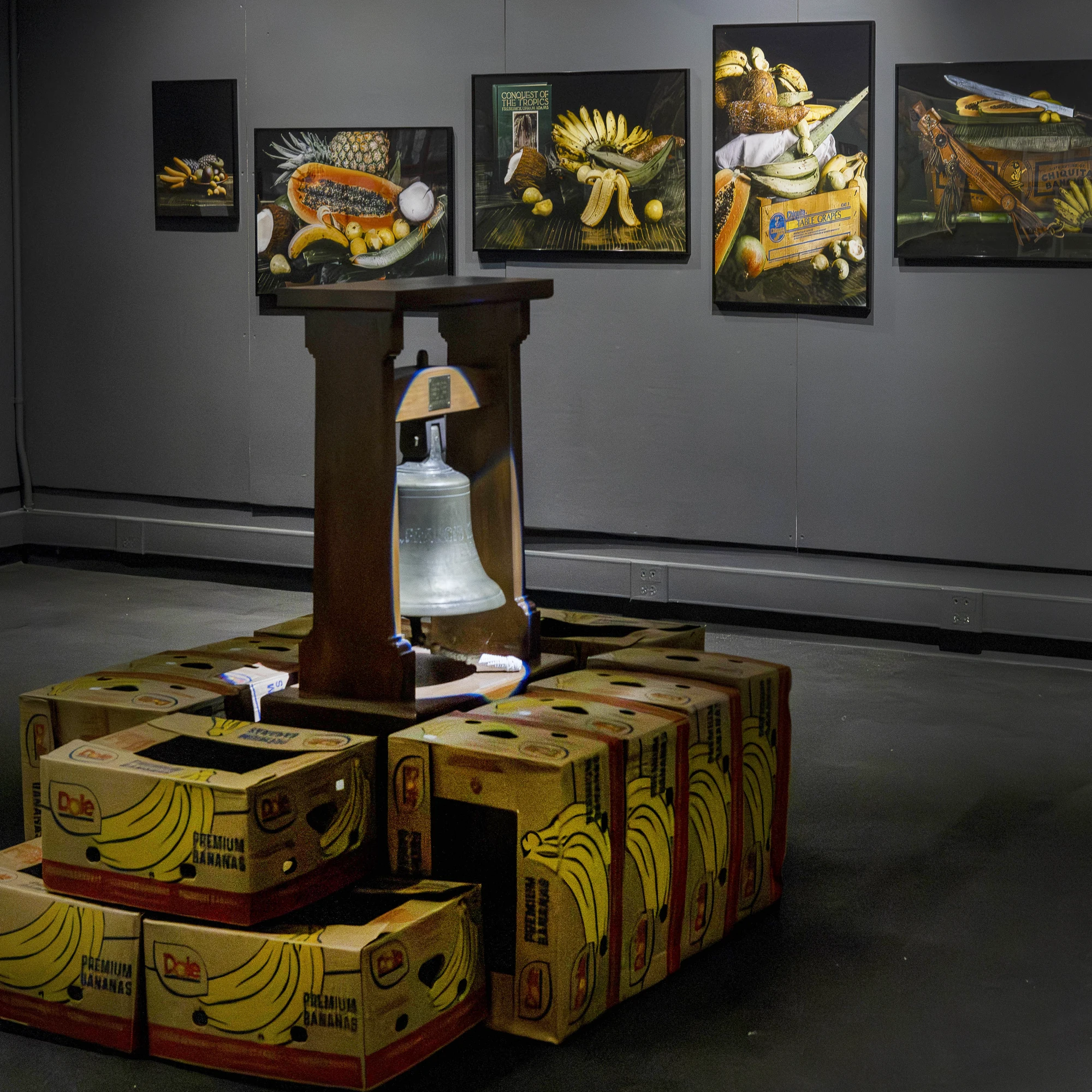 Bell from the Francis R Hart tanker sits atop banana boxes centered in a dark gallery with framed artifacts hanging on the wall behind.