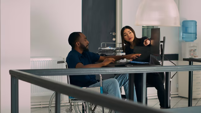 Two coworkers, one in a wheelchair, discussing work while pointing at a computer screen in an office.