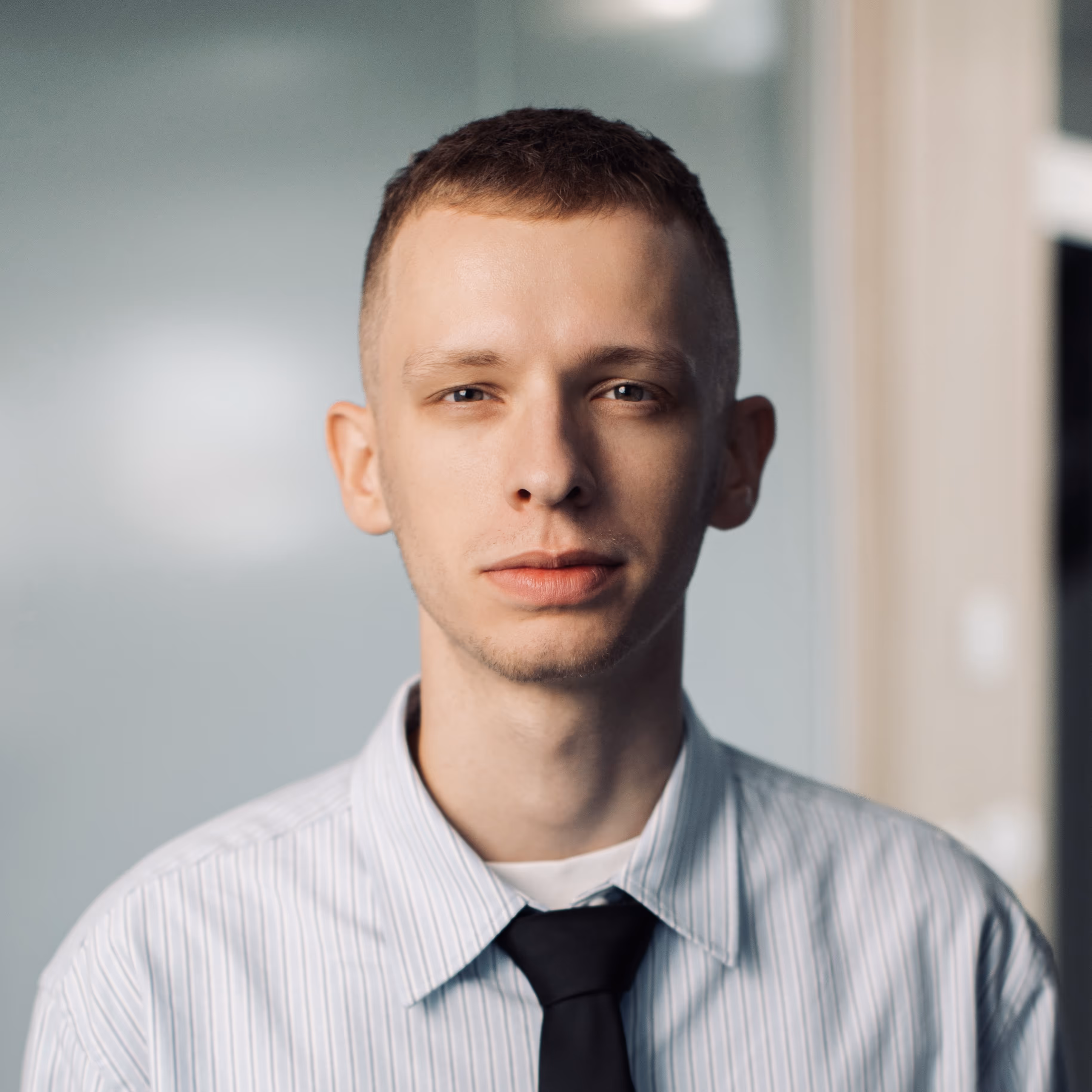 A young man with short dark hair and a neutral expression, wearing a light striped shirt and a black tie. The portrait is lit with a dramatic contrast, highlighting his face against an office background, creating a bold mood.