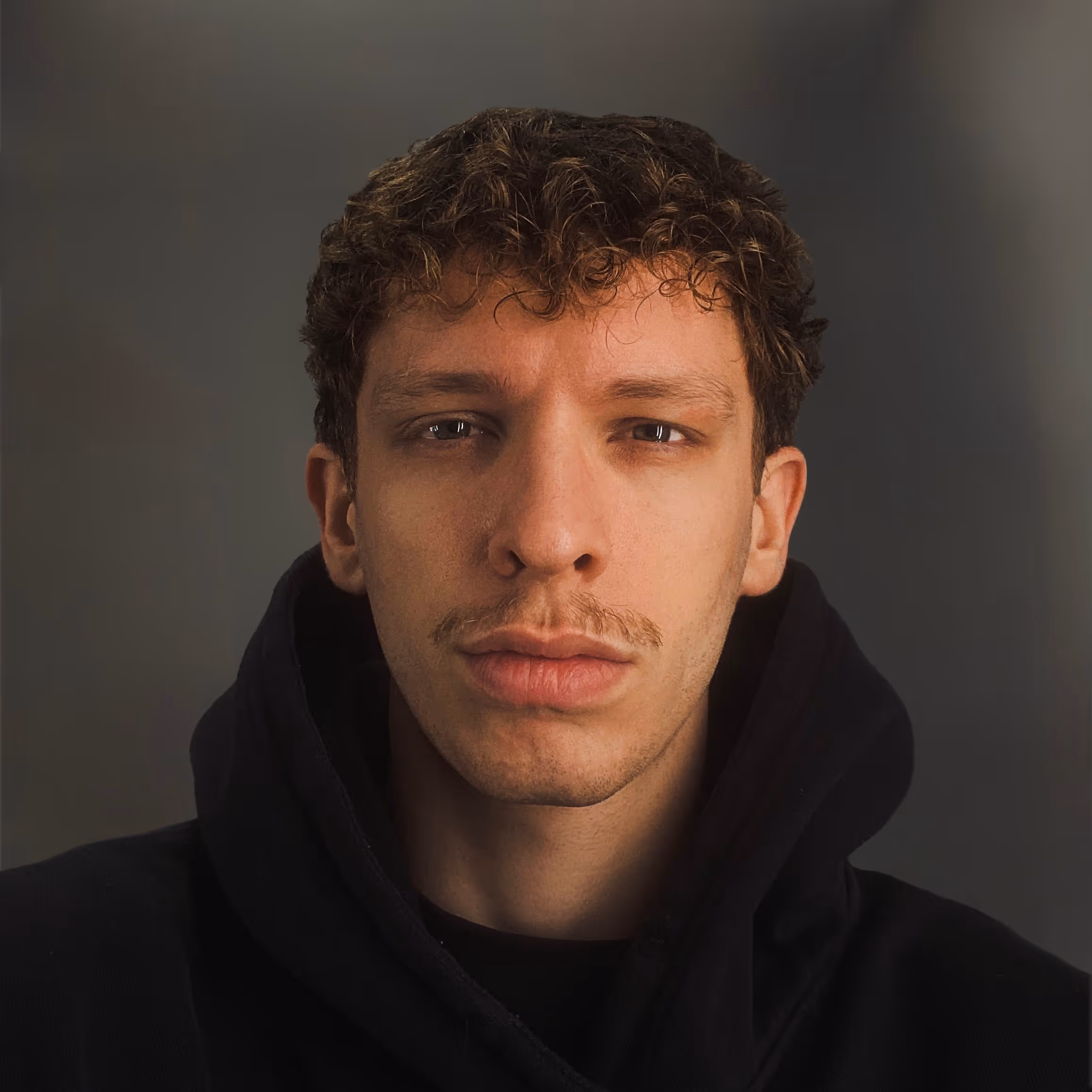 A young man with curly hair, dark mustache and a neutral expression, wearing a dark hoodie. The portrait is lit with a dramatic contrast, highlighting his face against a deep blurred background, creating a bold mood.