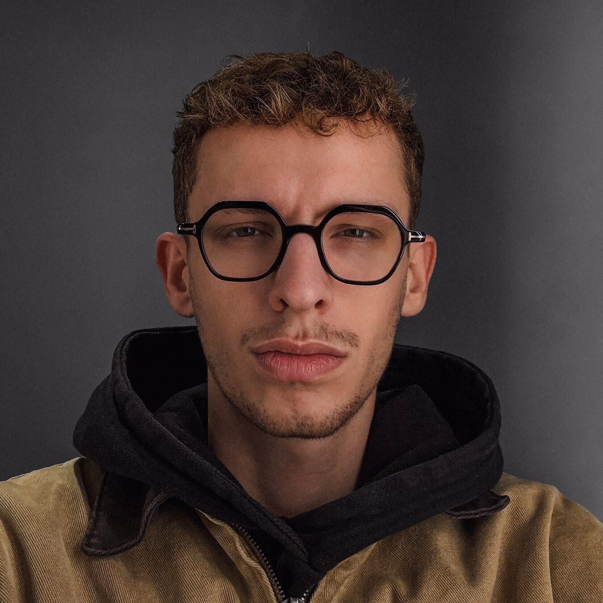 A young man with curly hair, black-framed glasses, a dark mustache, and a neutral expression, wearing a dark hoodie and a beige canvas jacket. The portrait is lit with dramatic contrast, highlighting his face against a deep, blurred background, which creates a bold mood.