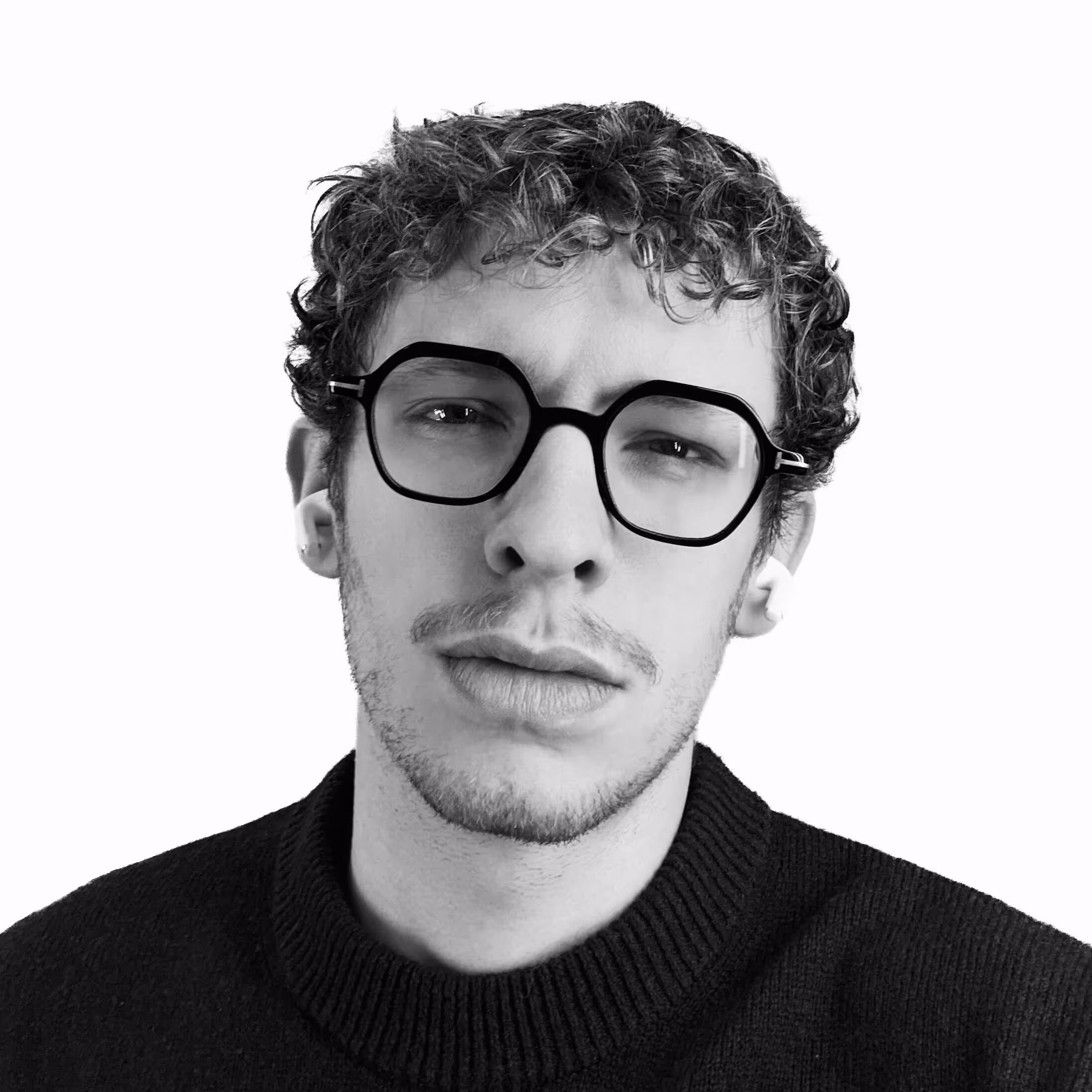 Young man with curly hair, black thick-rimmed glasses, and a black sweater, wearing white wireless earbuds against a white background.
