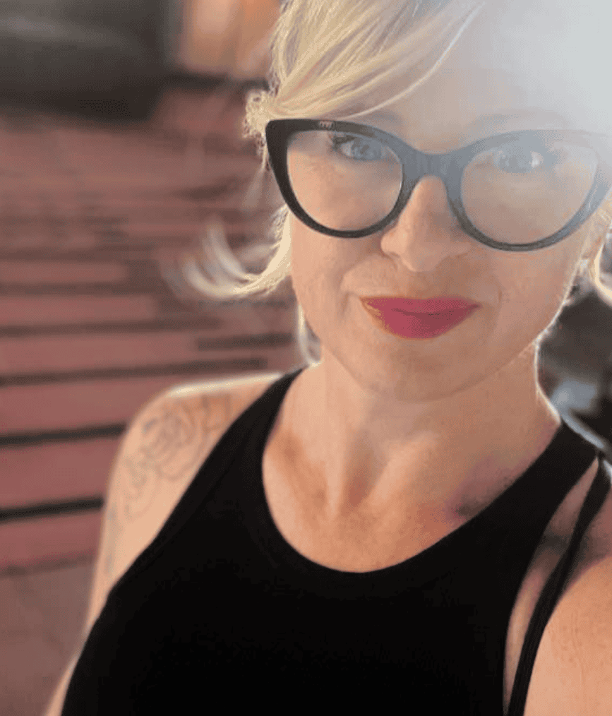 Outdoor portrait of Danielle Krysa wearing black-framed glasses and a sleeveless black top, photographed in warm light with a softly blurred urban background.