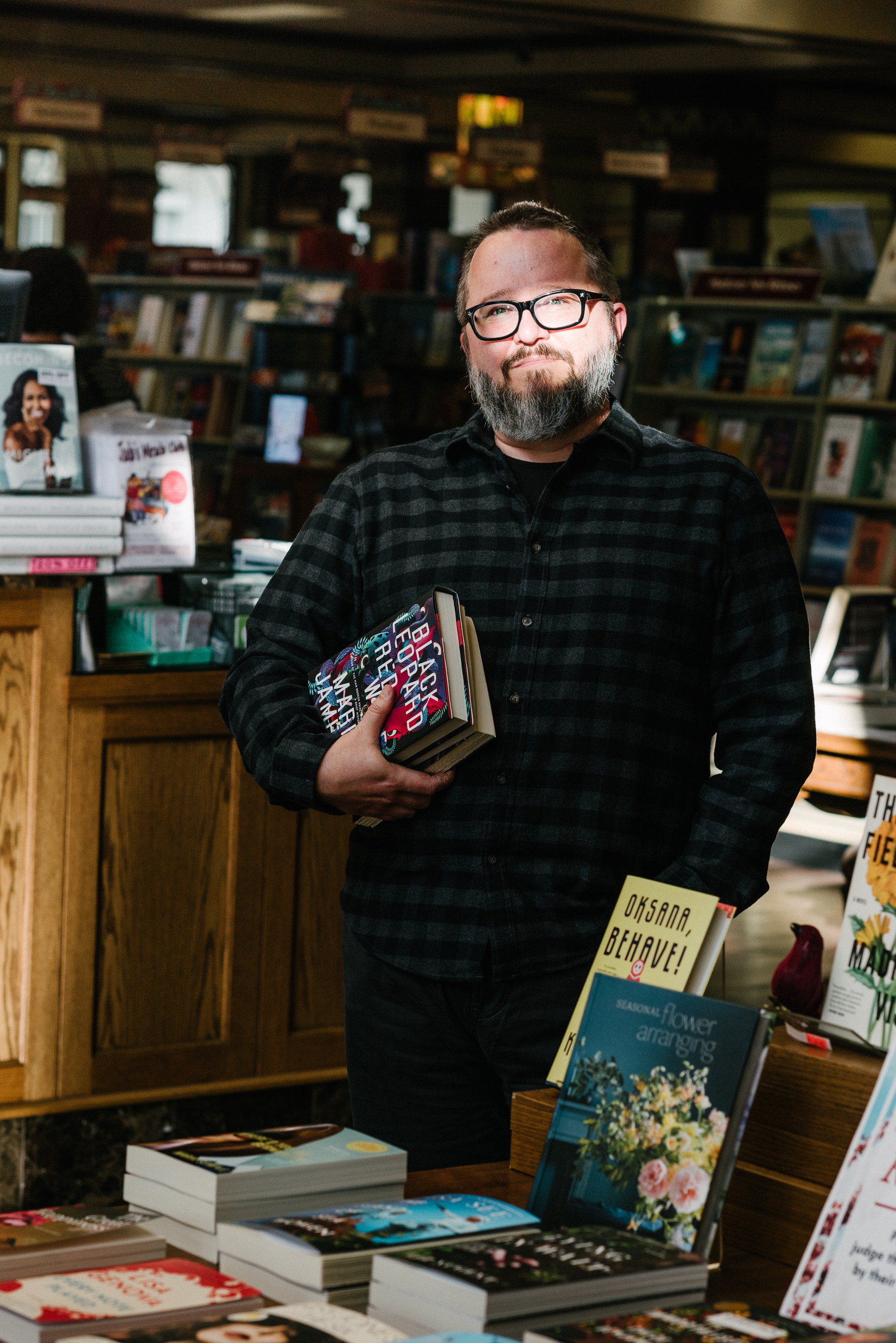 Portrait of Robert J. Wiersema standing in a bookstore, wearing glasses and a dark plaid shirt, holding a stack of books among shelves and displays.