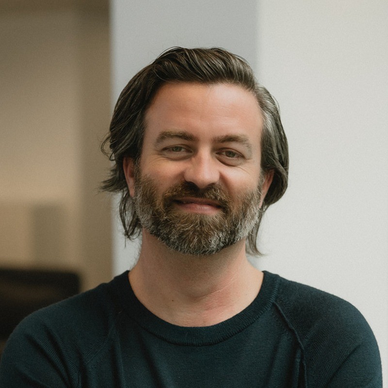 Indoor portrait of Casey Milone with a beard and medium-length dark hair, wearing a dark crewneck sweater and smiling slightly at the camera.