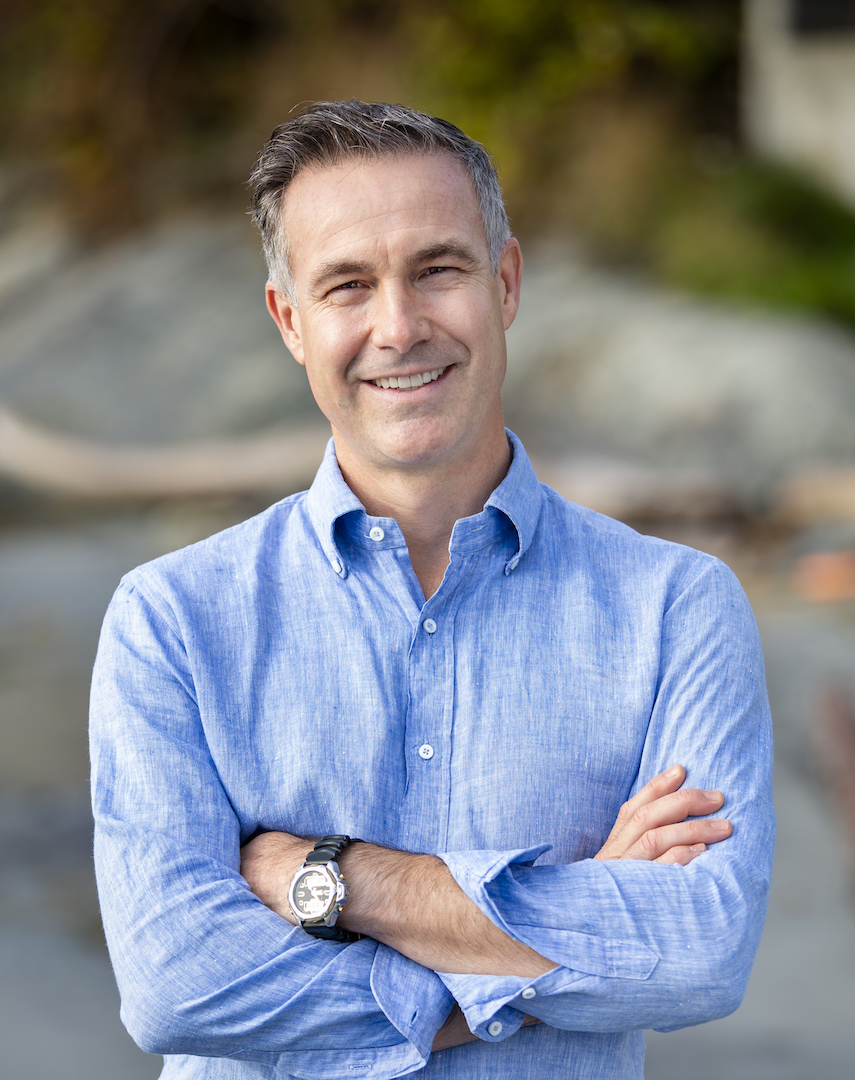 Outdoor portrait of Niobe wearing a light blue button-up shirt with rolled sleeves, arms crossed, smiling at the camera with a softly blurred natural background.