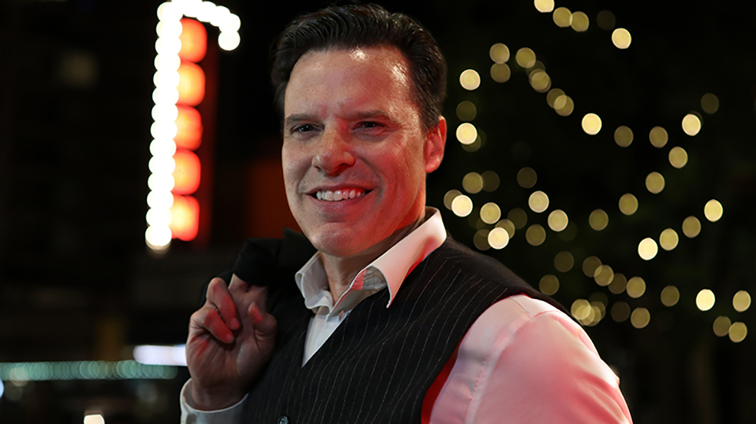 Nighttime portrait of Johnathan Henevey wearing a white shirt and dark vest, smiling while holding a jacket over his shoulder, with softly blurred lights in the background.