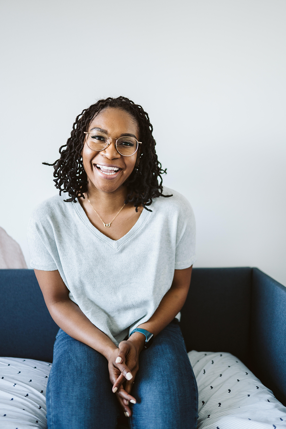 Portrait of Sharon Milone seated on a couch, smiling warmly at the camera, wearing glasses, a light grey top, and jeans against a clean, light background.