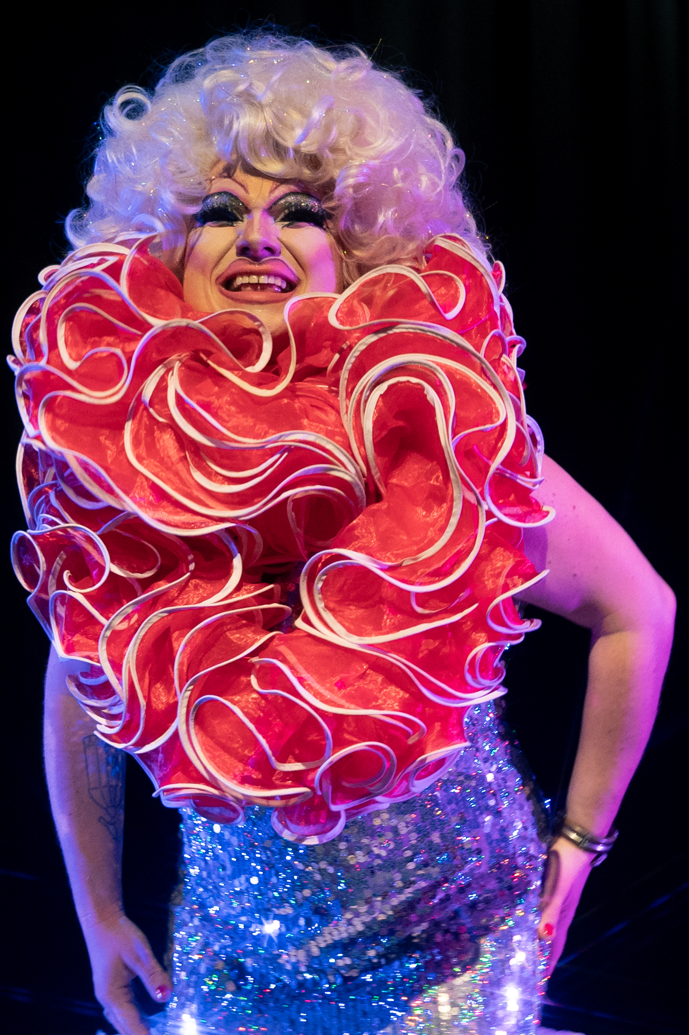 A drag performer on stage wearing a dramatic red ruffled collar, glittering blue sequined outfit, and a curly white wig, smiling under colourful stage lighting against a dark background.