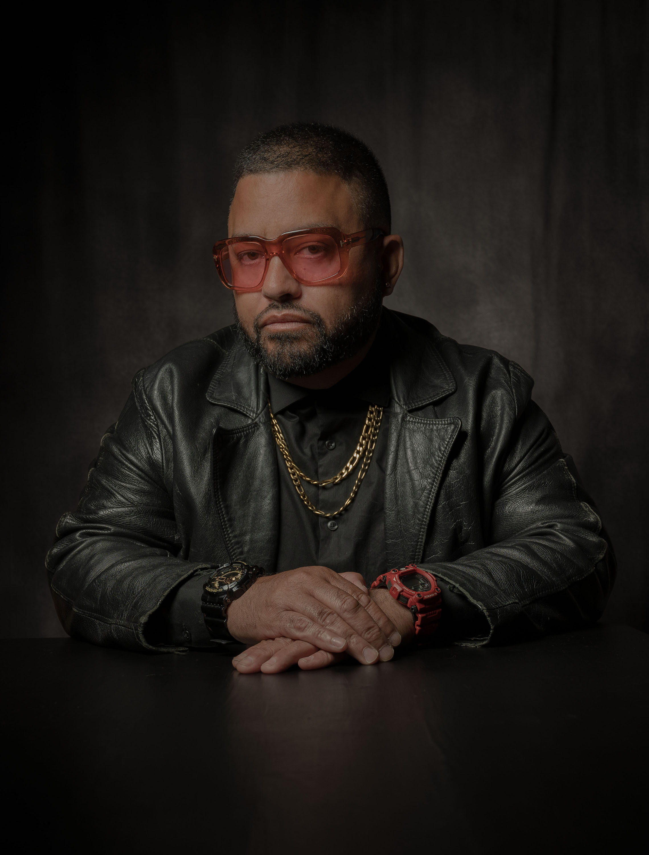 Studio portrait of a person wearing red-tinted glasses, a black leather jacket, and gold chains, seated at a table against a dark backdrop.
