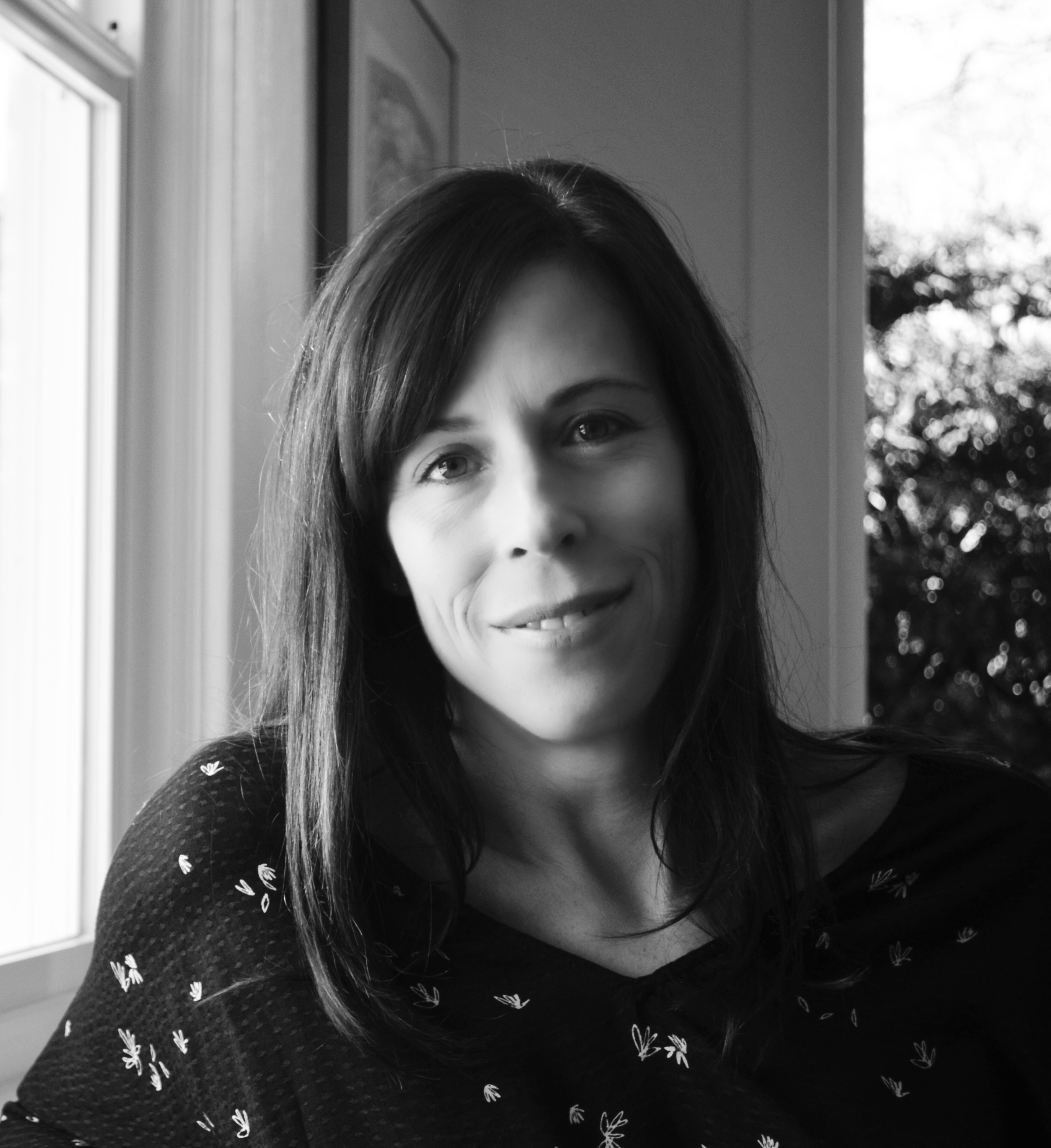 Black and white portrait of a person with shoulder-length dark hair, seated indoors near a bright window, looking calmly toward the camera.