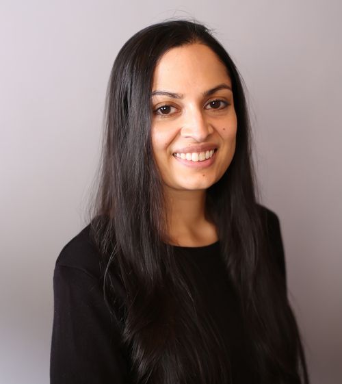 Headshot of Canadian author Shashi Bhat, smiling, with long dark hair, wearing a black top against a grey background.