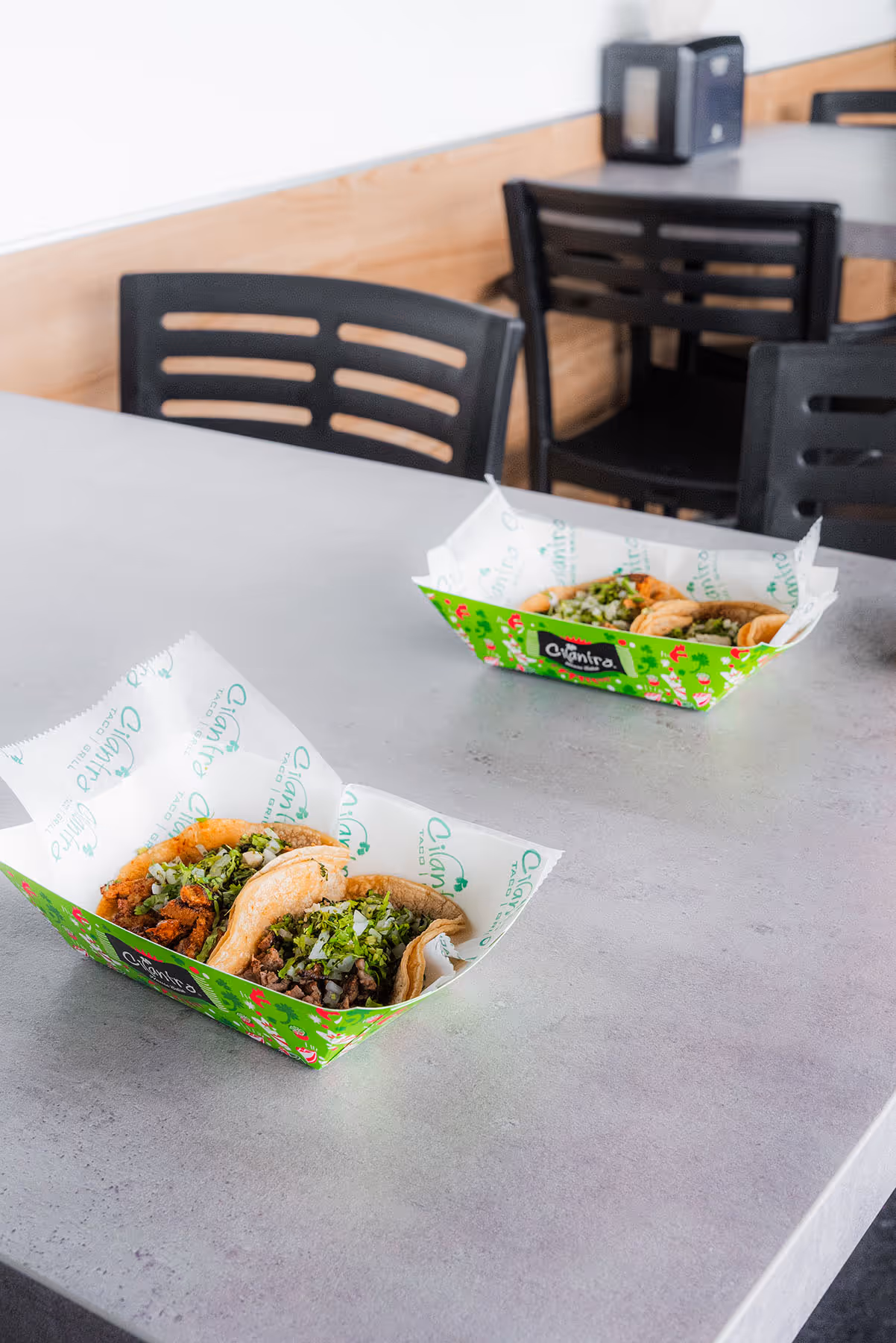 Two 'Cilantro Taco Grill' cardboard trays on a gray table. Each tray holds two tacos served with meat, cilantro, and onion. The interior of a restaurant is visible in the background.