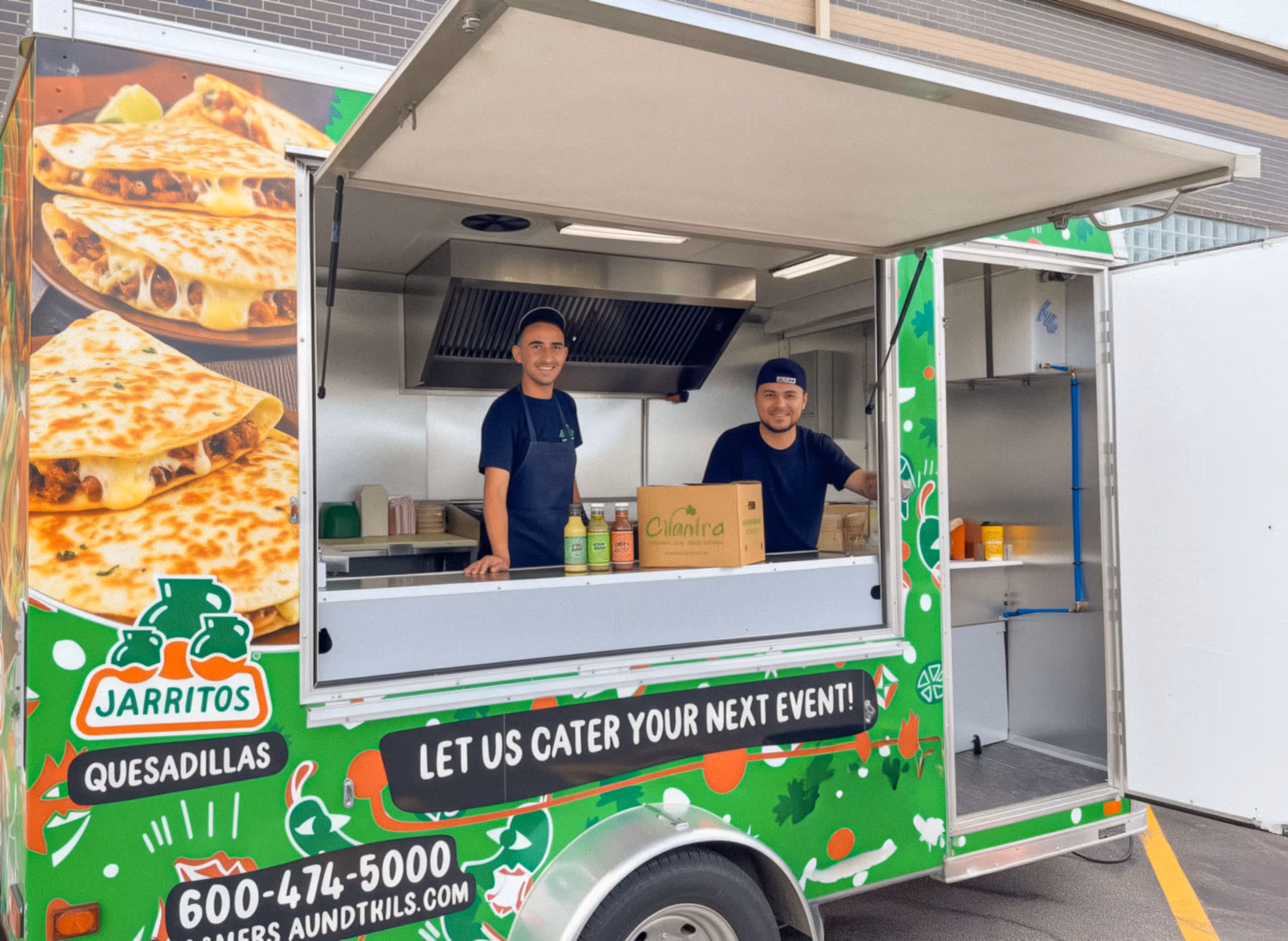 Two friendly caterers smiling from the service window of the green 'Cilantro' food truck. The truck advertises 'Let Us Cater Your Next Event!' and features Jarritos branding and delicious pictures of quesadillas.