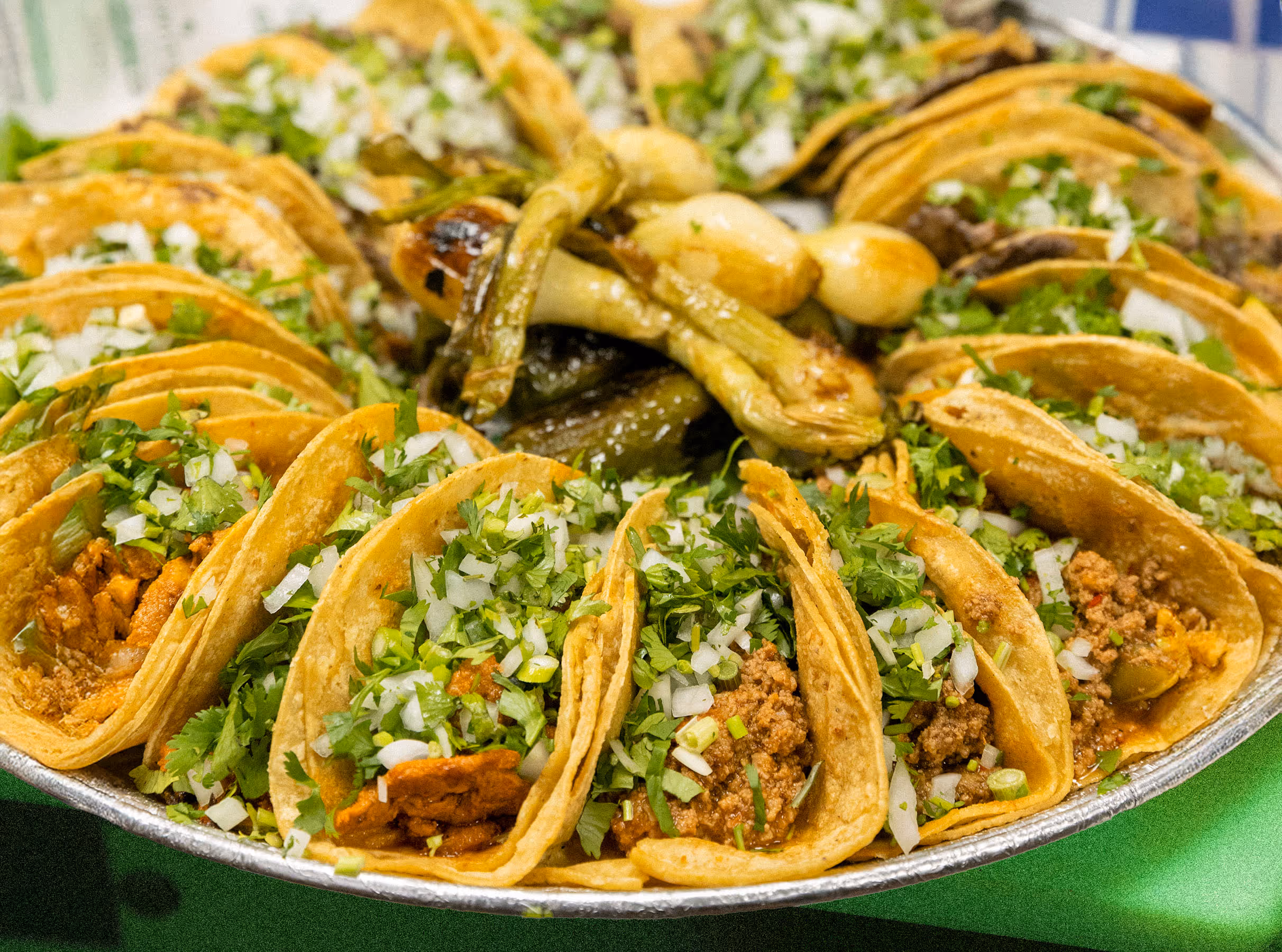 A large catering platter filled with various authentic Mexican street tacos, garnished with fresh cilantro and diced onions, accompanied by grilled green onions and jalapeños.