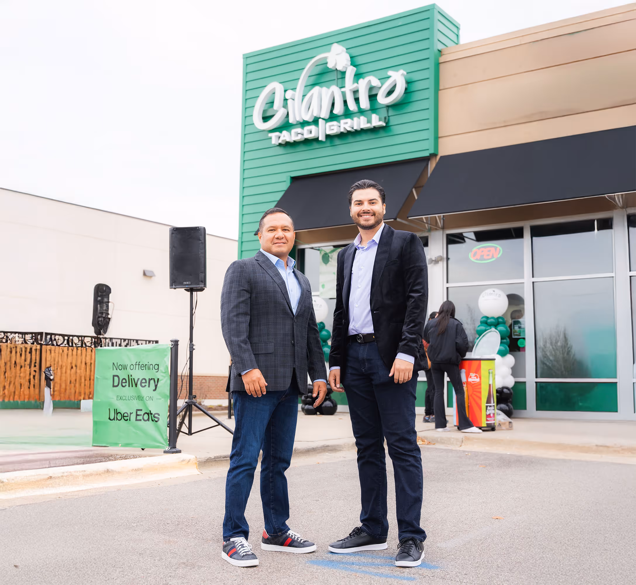 Two men, a business owner and a partner, pose confidently in front of a Cilantro Taco Grill restaurant with its distinctive green facade. A sign promoting Uber Eats delivery stands to the left.