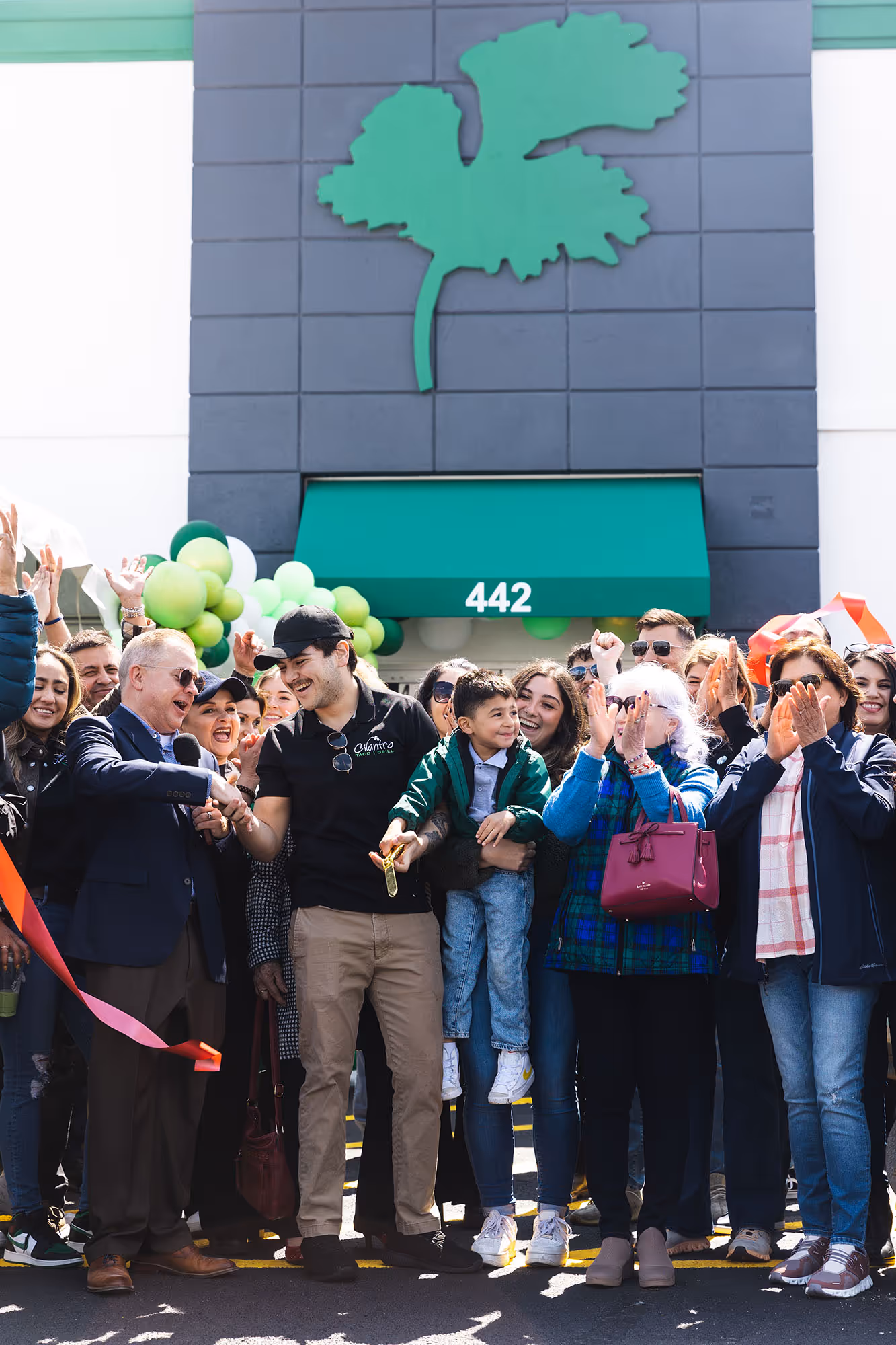 A crowd of people, including families and what appears to be a Mariachi band, celebrate a grand opening. A man in the center holds a young child while others clap and cheer, with a large green cilantro logo visible on the building behind them.