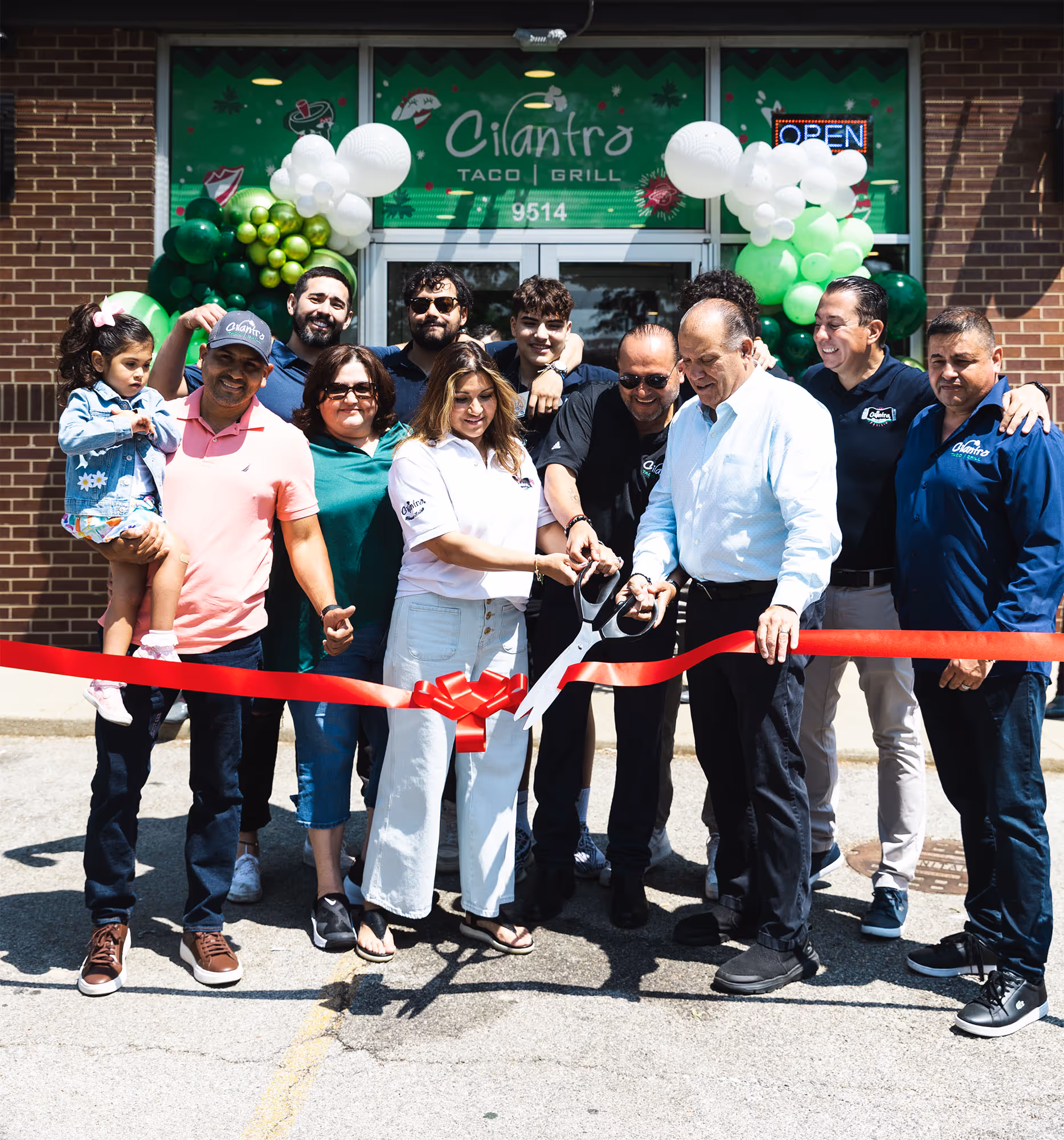 A large family and staff gather outside a Cilantro Taco Grill location, cutting a red ribbon with a pair of large scissors to signify a grand opening. Green and white balloons decorate the restaurant's entrance.