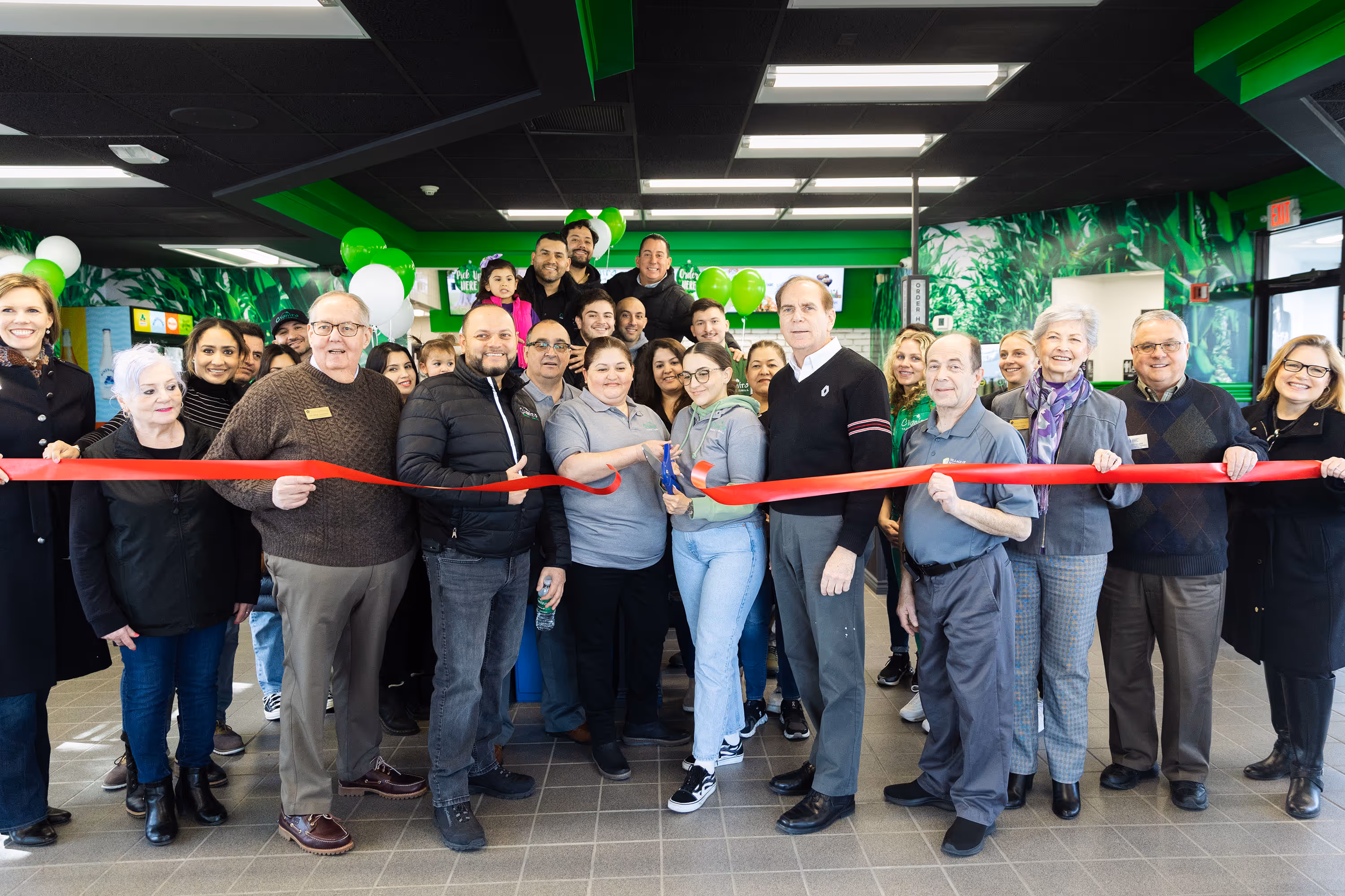 A large, diverse group of people, including family members, business partners, and community members, pose for a photo inside a restaurant, holding a red ribbon during a ribbon-cutting ceremony. The restaurant's interior is decorated with green, white, and black accents.