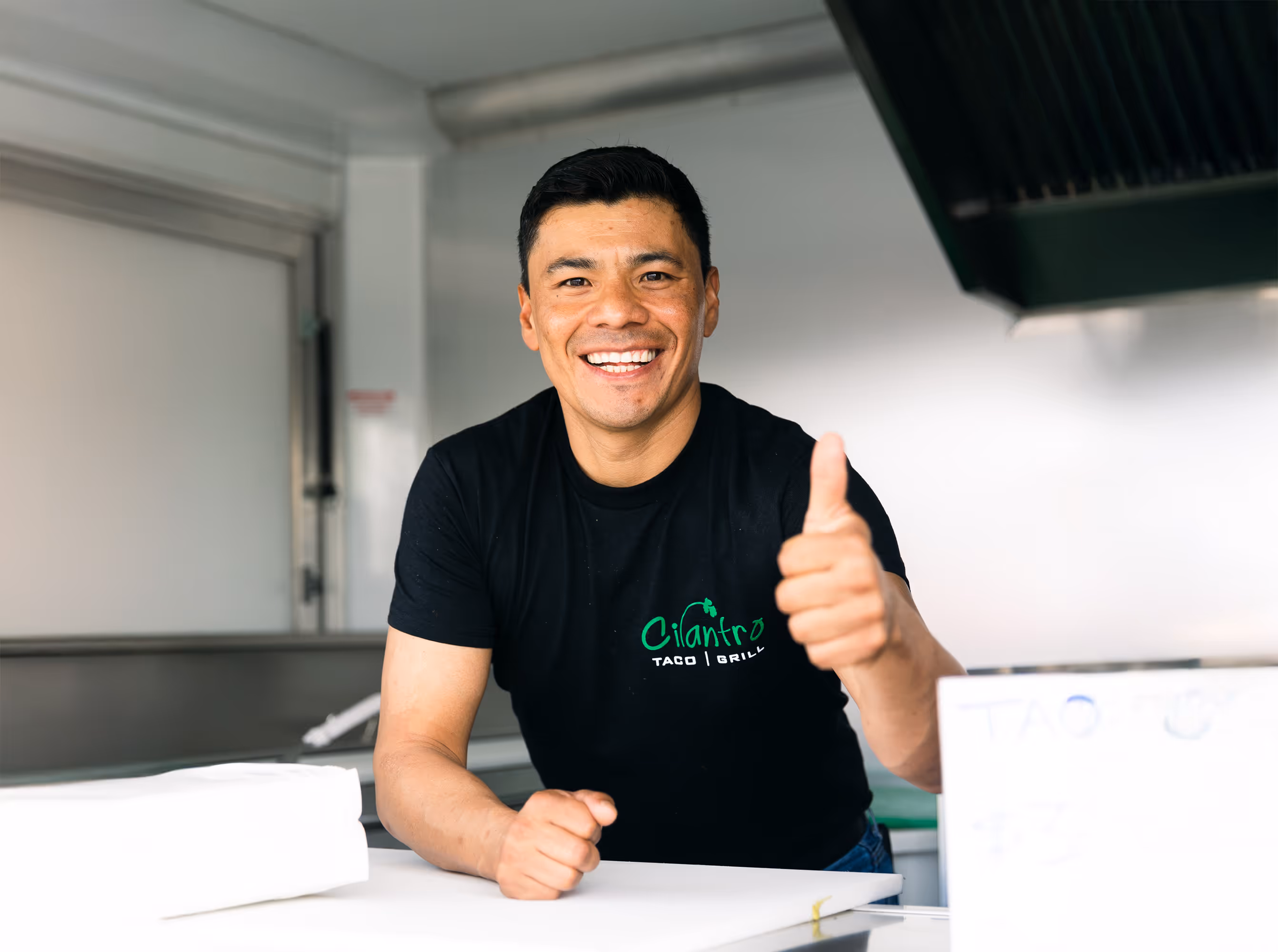 A male employee wearing a black t-shirt gives a thumbs-up and smiles brightly while standing behind a counter.