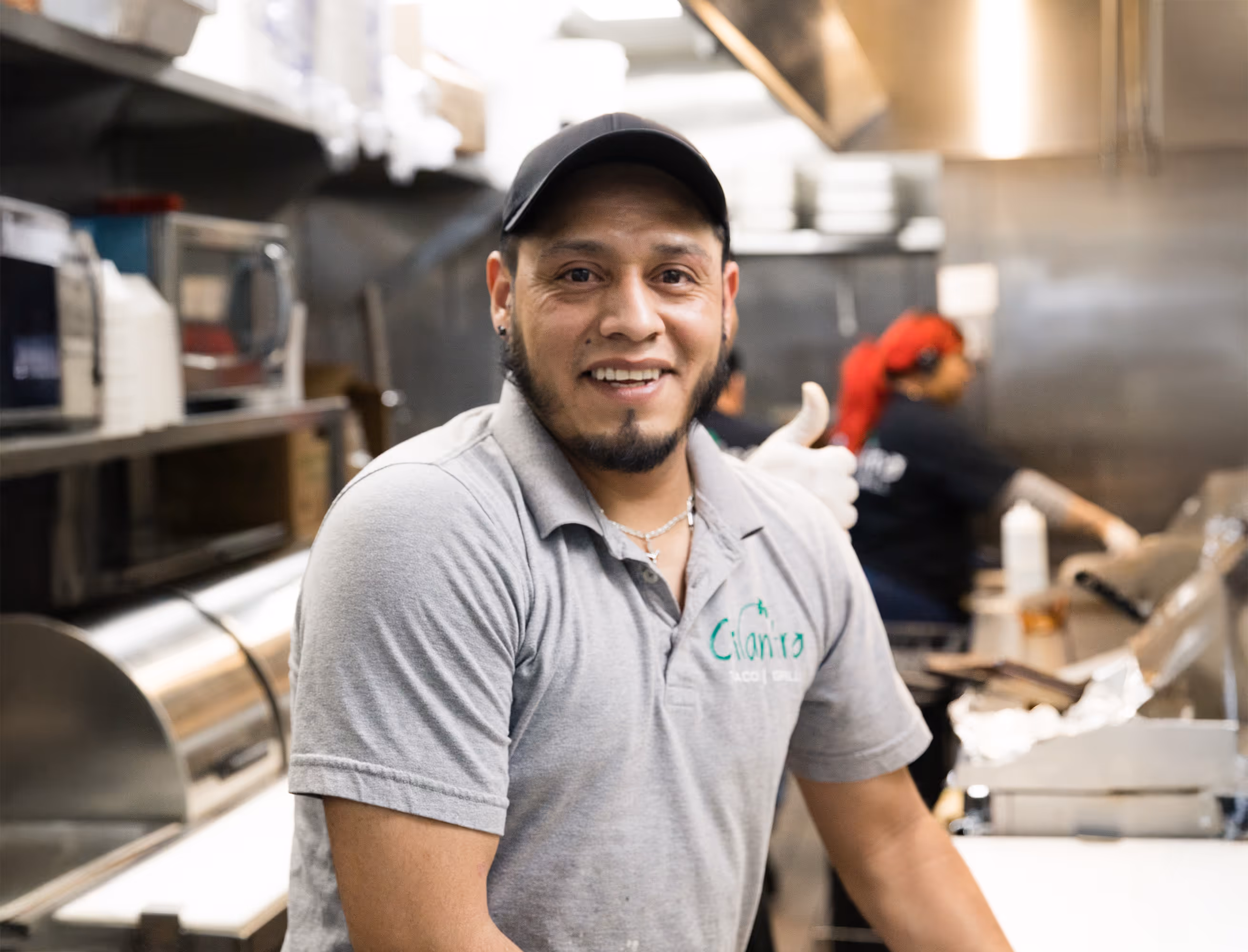 A male cook with a beard and a baseball cap smiles at the camera, giving a thumbs-up while working in a kitchen with another staff member visible in the background.