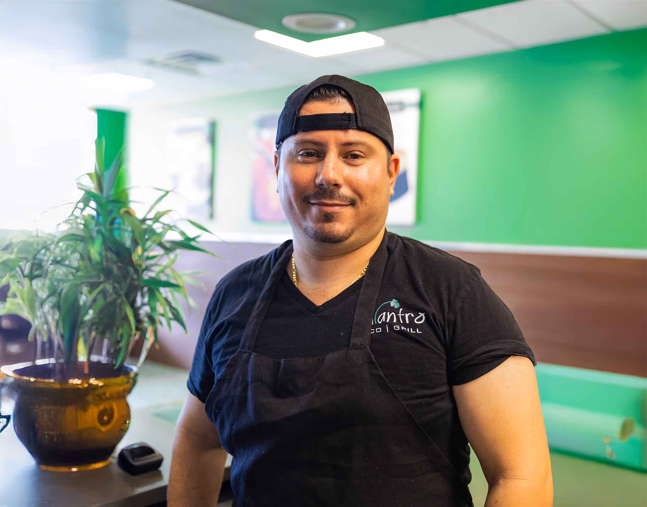 A male staff member with a mustache, beard, and backward baseball cap smiles and poses for a portrait inside the restaurant with green walls and a potted plant nearby.
