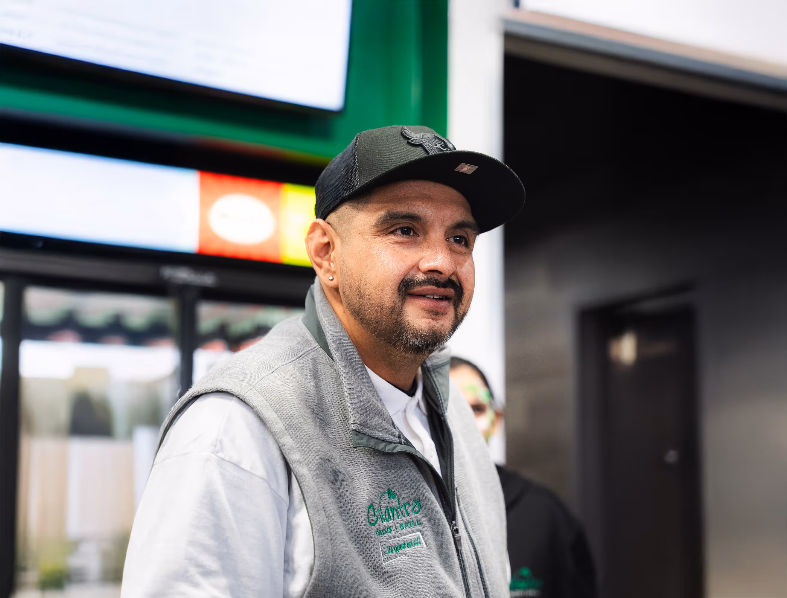 A male staff member with a goatee and a Bulls cap smiles while wearing a gray fleece vest embroidered with the "Cilantro Taco Grill" logo.