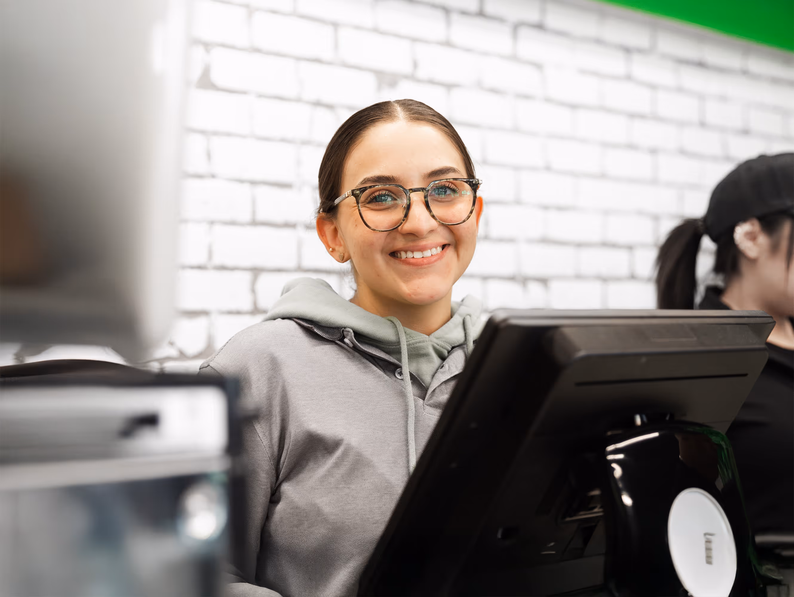 A young female employee with glasses smiles warmly as she stands behind the counter, with a white brick wall in the background.