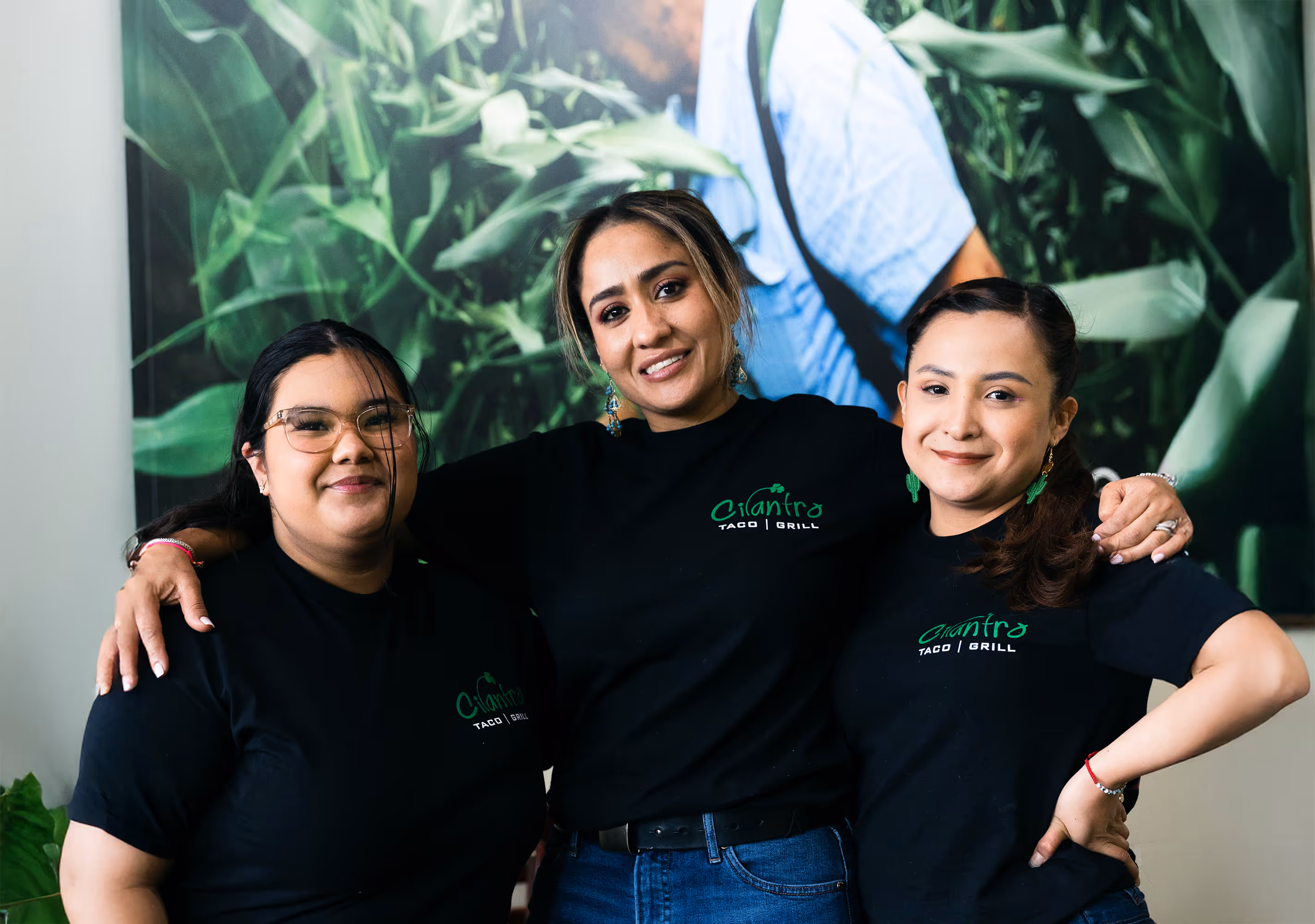Three smiling female employees in black t-shirts pose together with their arms around each other's shoulders in front of a large mural.