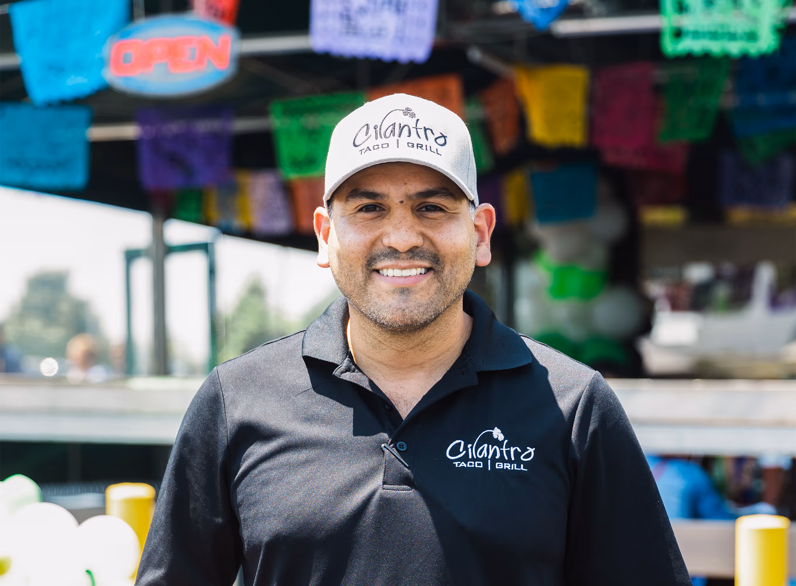 A smiling male staff member with a goatee and a gray hat stands in front of colorful papel picado banners, wearing a black polo shirt with the "Cilantro Taco Grill" logo.