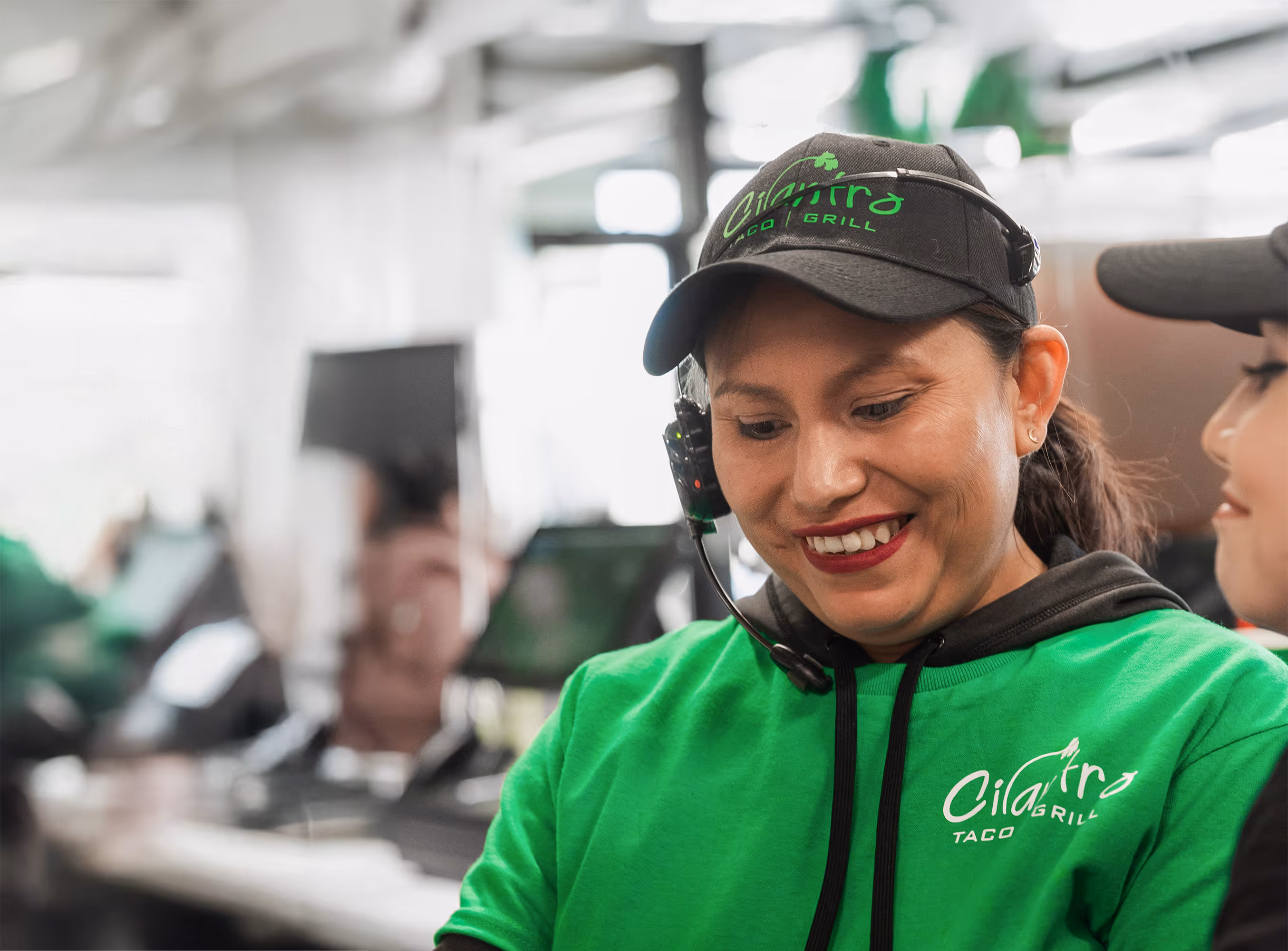 A female employee with a headset and a bright green hoodie smiles while working at the counter.