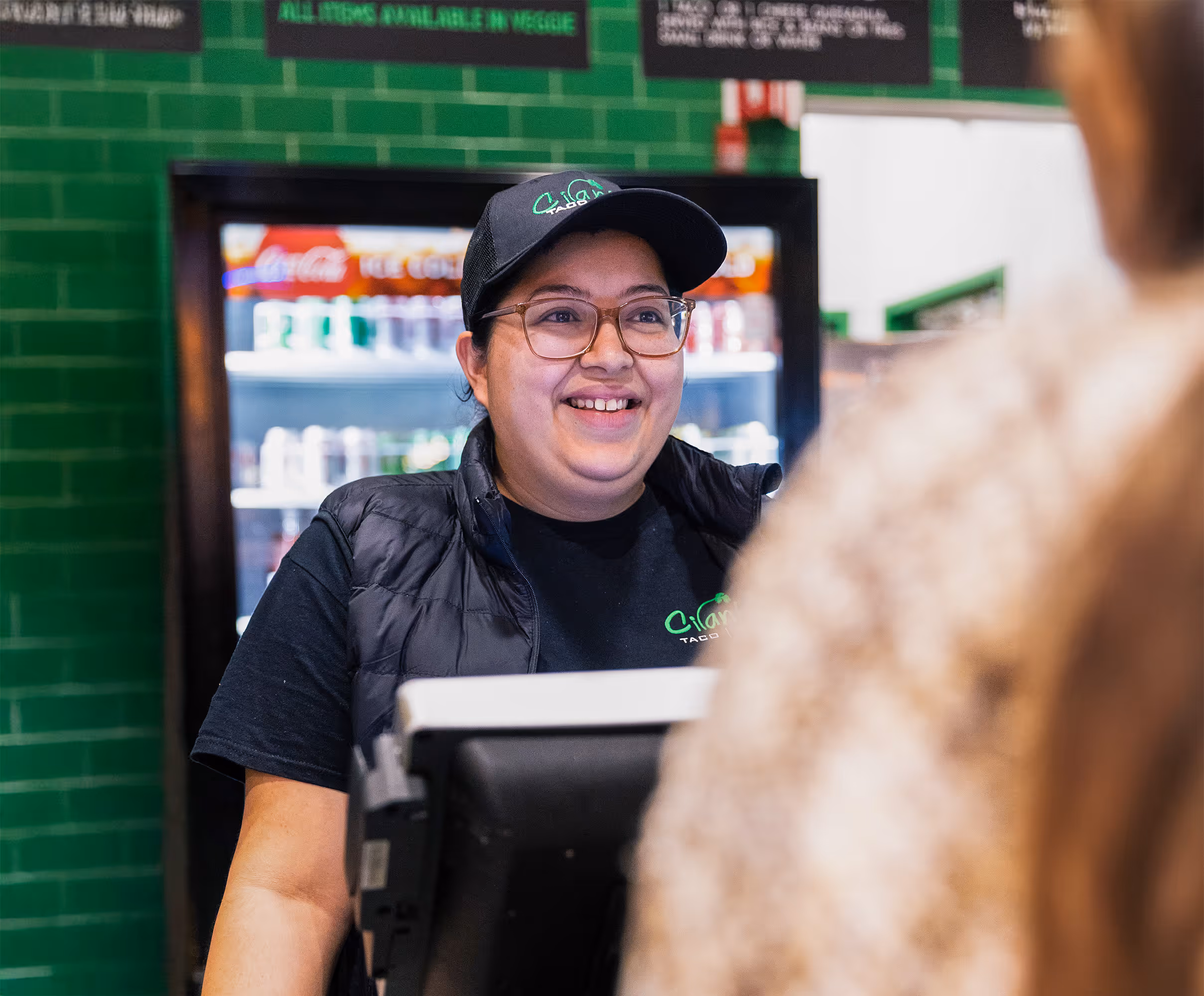 A female employee with a hat and glasses smiles warmly at a customer from behind the counter, with a refrigerated drink display in the background.