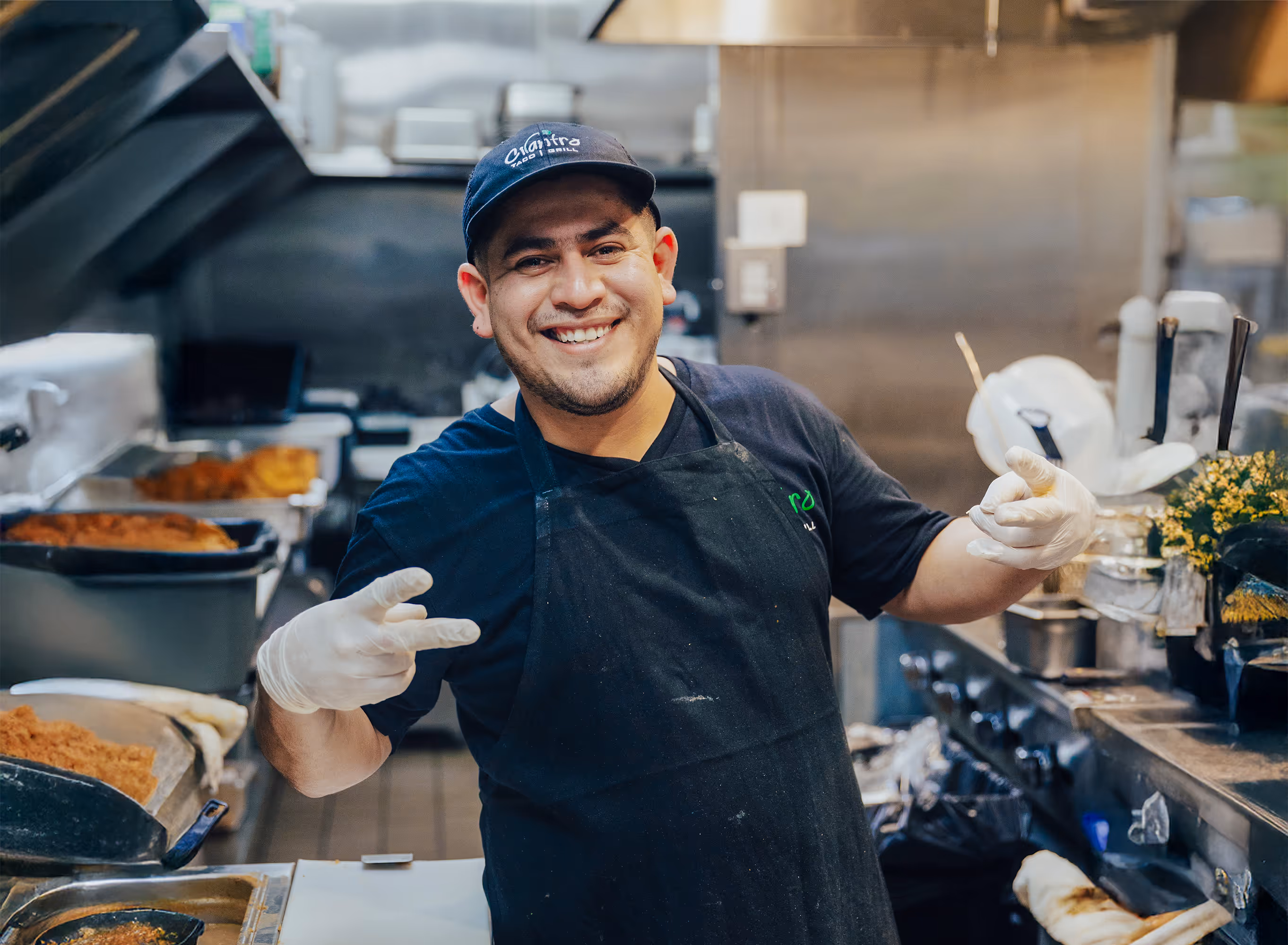 A smiling male chef in a black apron and gloves gives two peace signs to the camera from inside a professional kitchen.