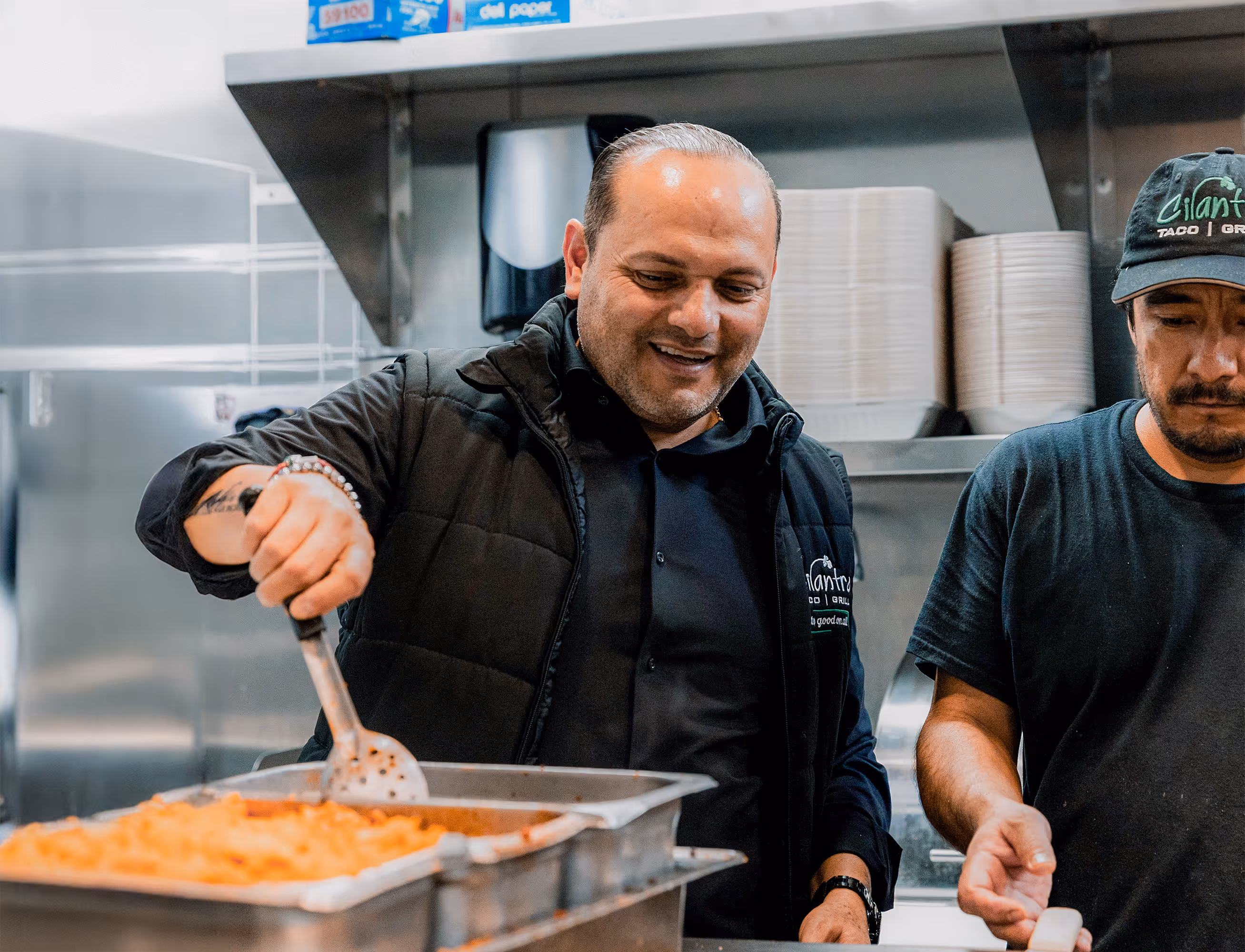 A male manager wearing a black vest smiles while using a large metal spatula to stir a tray of food in the kitchen.