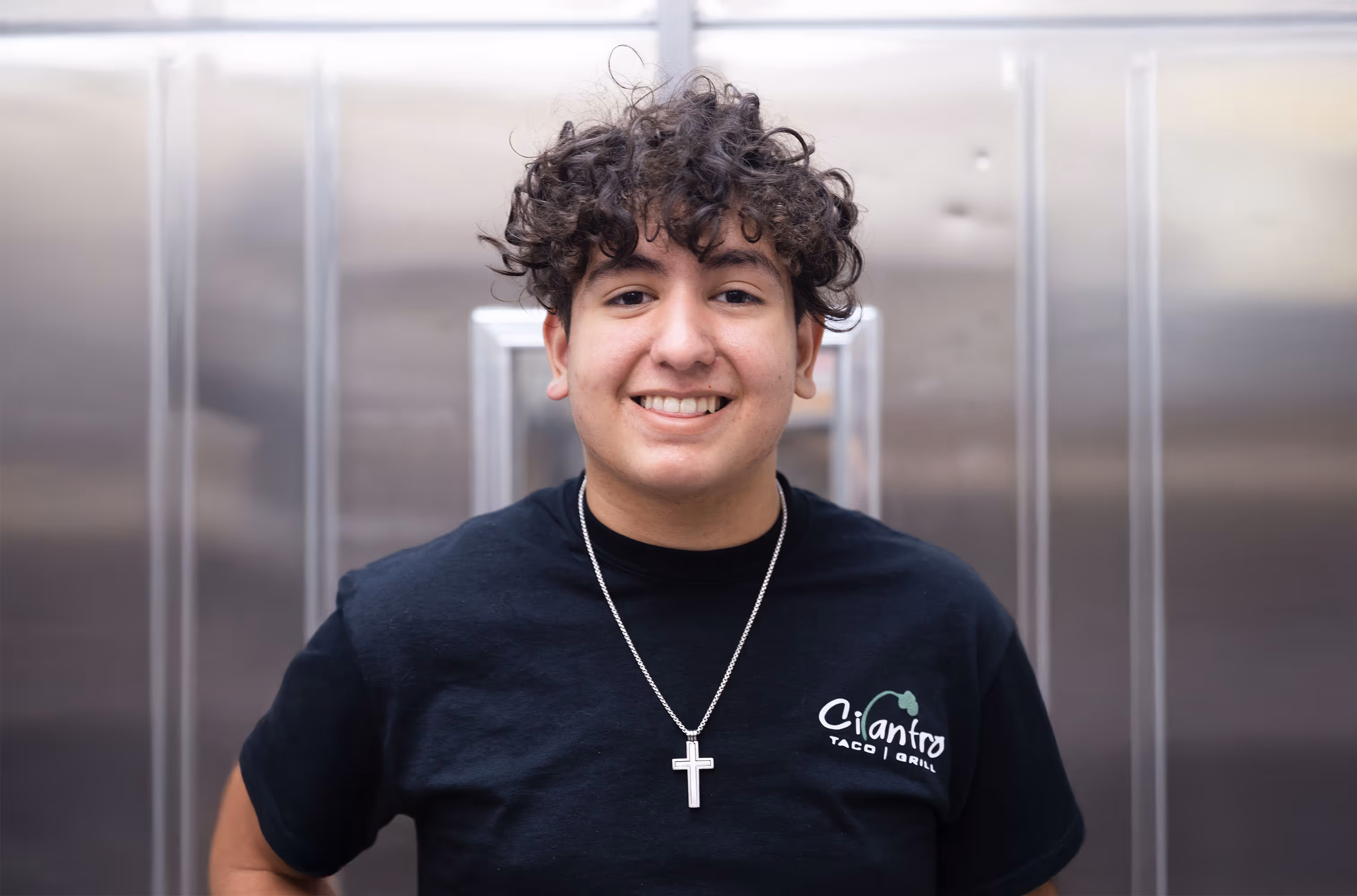A smiling male employee with curly hair and a silver cross necklace stands in front of a stainless steel kitchen door, wearing a black Cilantro Taco Grill t-shirt.