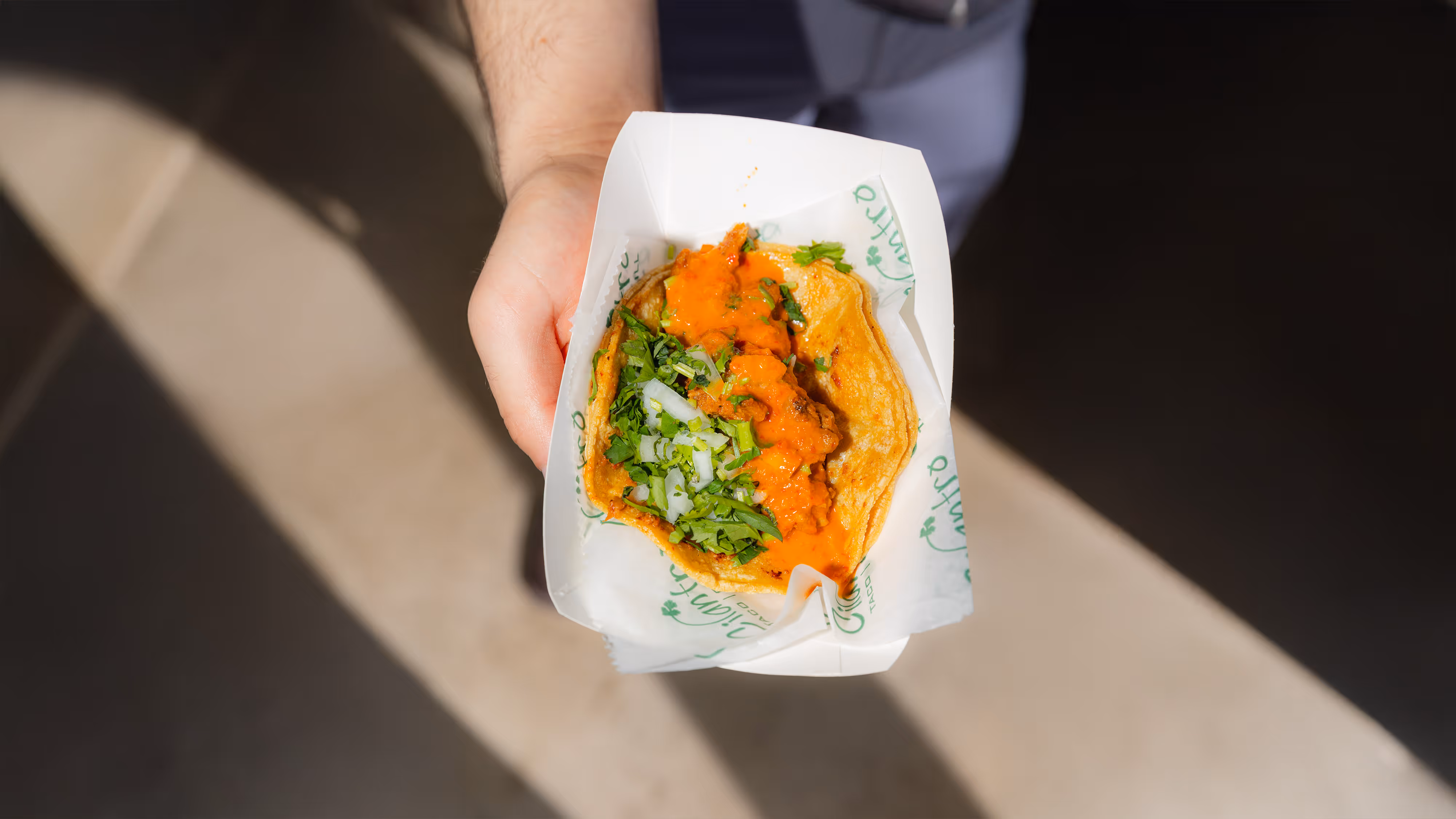 Hand holding a taco with meat, chopped onions, cilantro, and orange salsa in a paper tray.