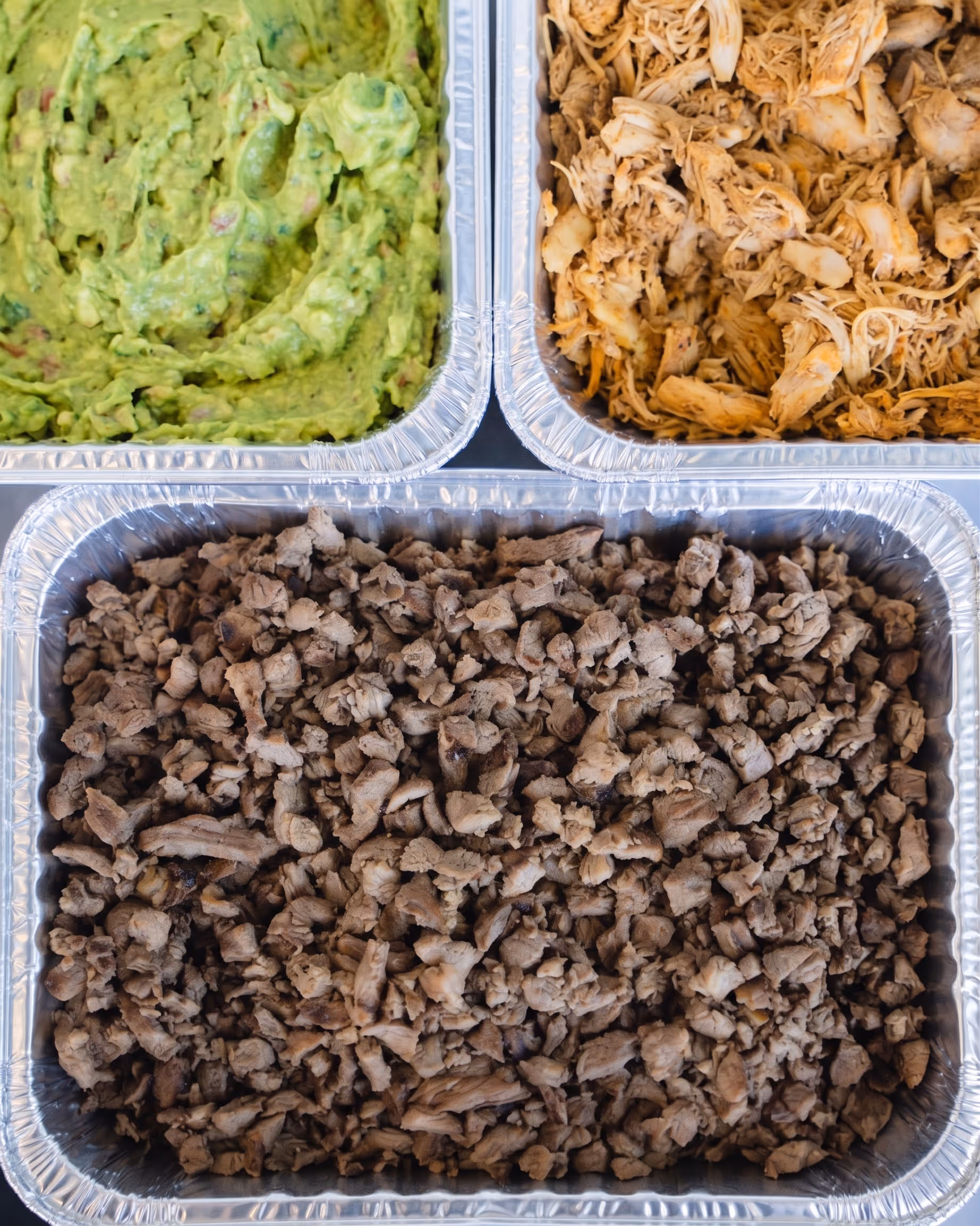 Aluminum trays with cooked shredded chicken, seasoned beef, and guacamole for a taco bar.