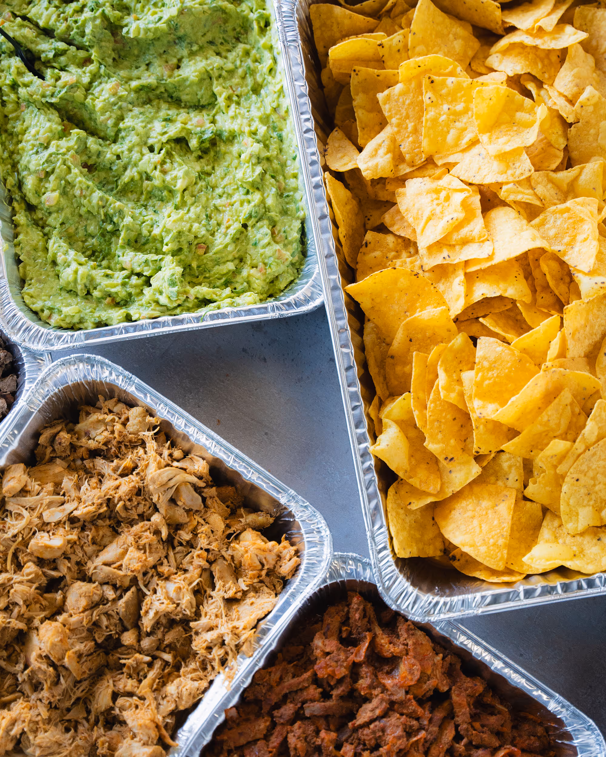 Aluminum trays with guacamole, tortilla chips, shredded chicken, and seasoned beef for a taco bar.