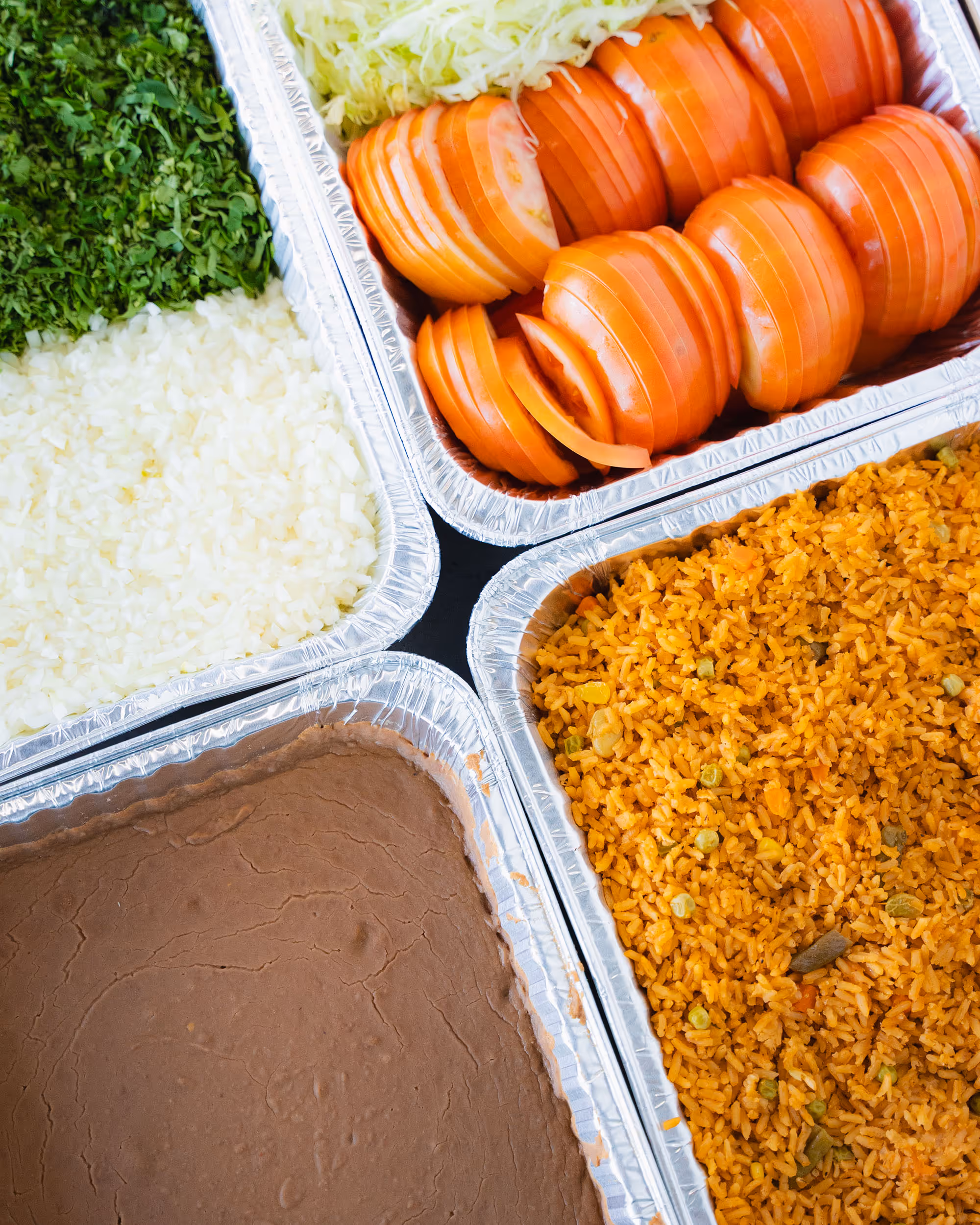 Aluminum trays with Mexican rice, refried beans, sliced tomatoes, chopped onions, cilantro, and shredded lettuce.