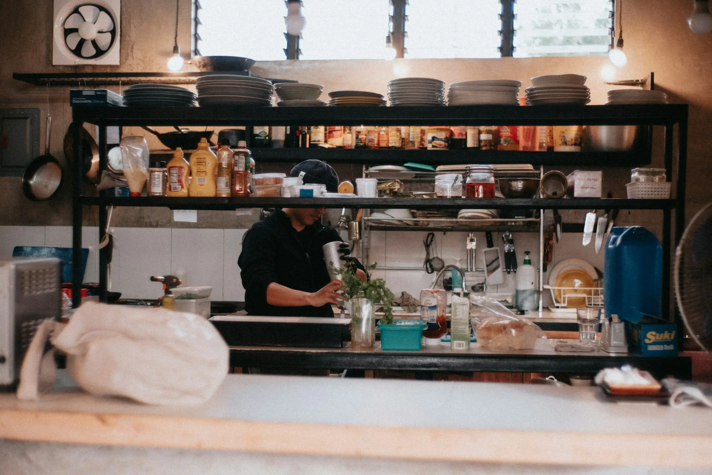 Person preparing food behind a kitchen counter with shelves holding plates, condiments, and kitchen tools.