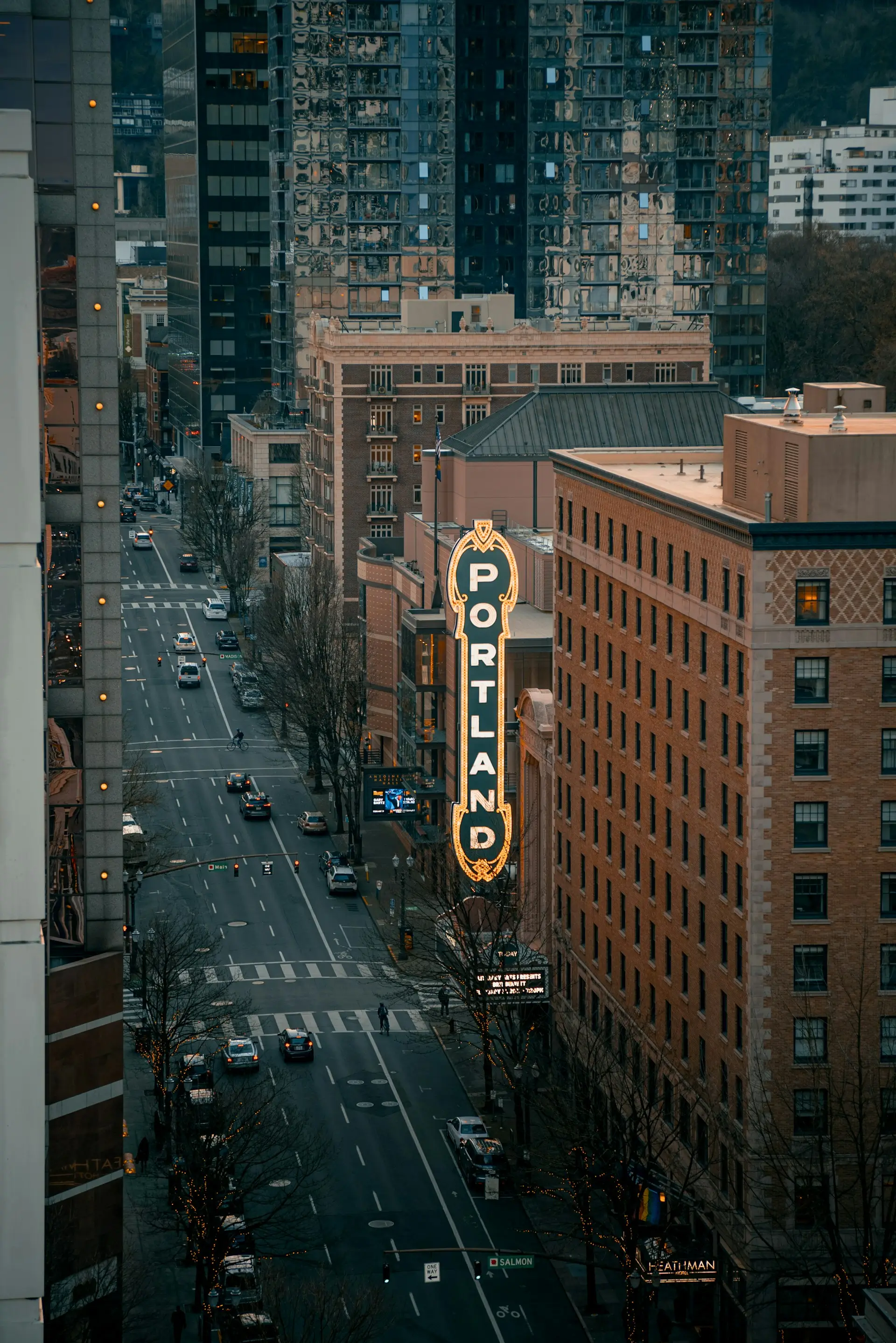 Downtown Portland street view with illuminated vertical 'PORTLAND' sign on a building and several cars on the road at dusk.