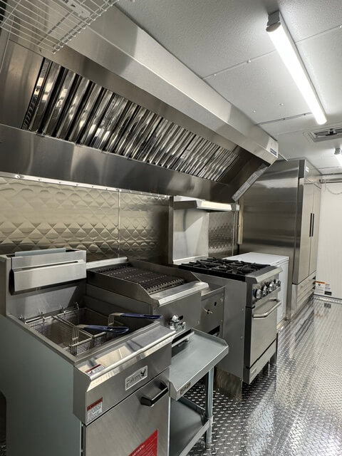Stainless steel commercial kitchen appliances including a deep fryer, grill, stove, and a refrigeration unit with overhead ventilation.