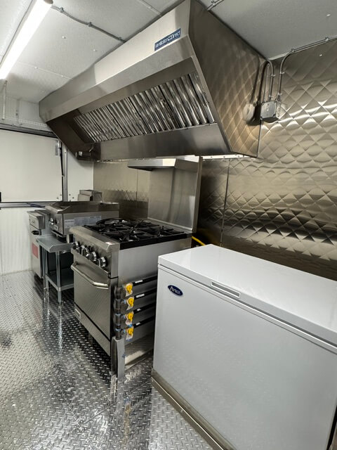 Interior of a commercial kitchen featuring a stainless steel stove with four burners, a large exhaust hood, and a white chest freezer on a diamond plate floor.