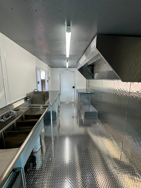 Interior of a stainless steel mobile commercial kitchen with sinks, countertops, and ventilation hood.